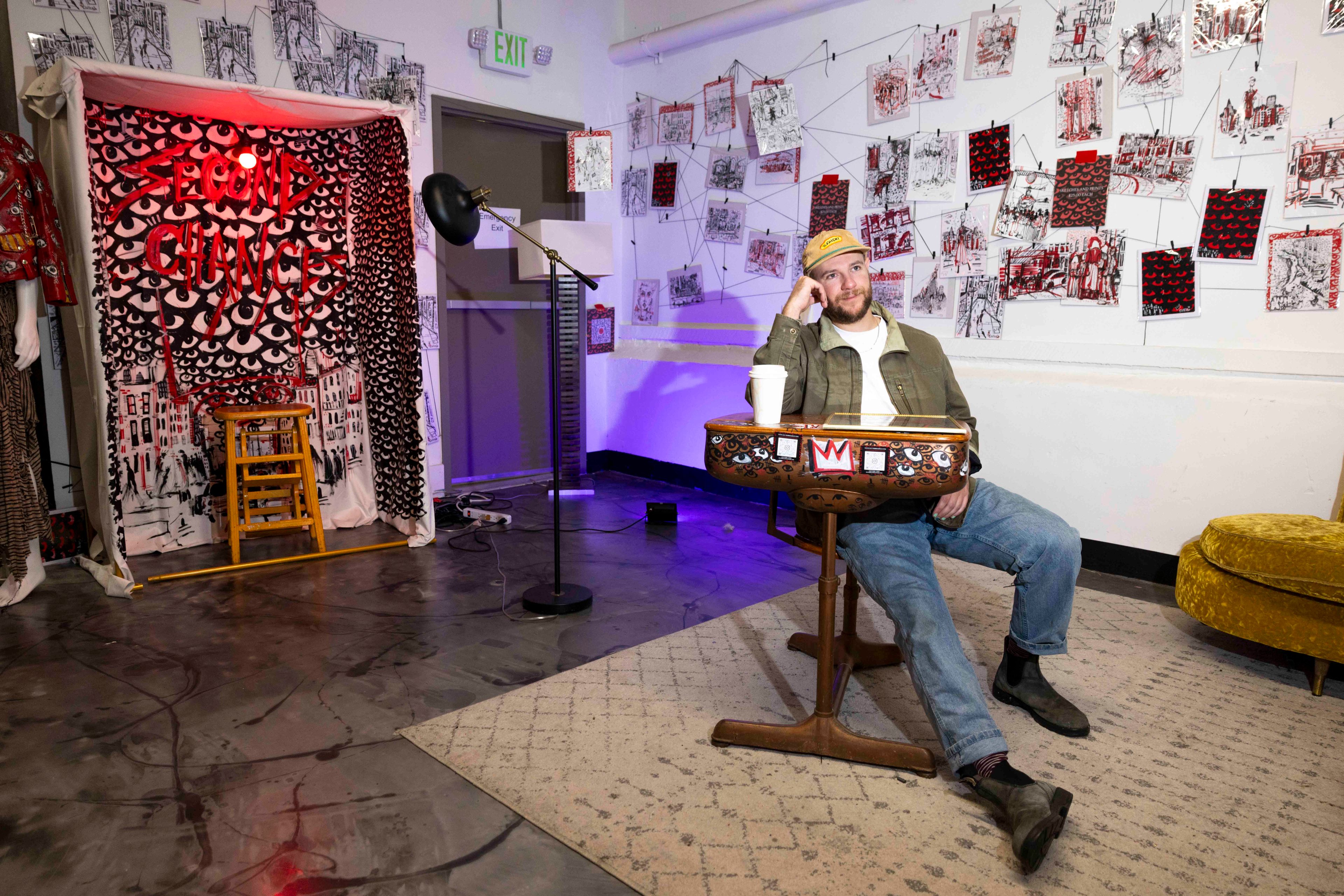 A man sits at a decorated table in an art-filled room, surrounded by sketches and colorful lights. A small stage with "Second Chance" is nearby.