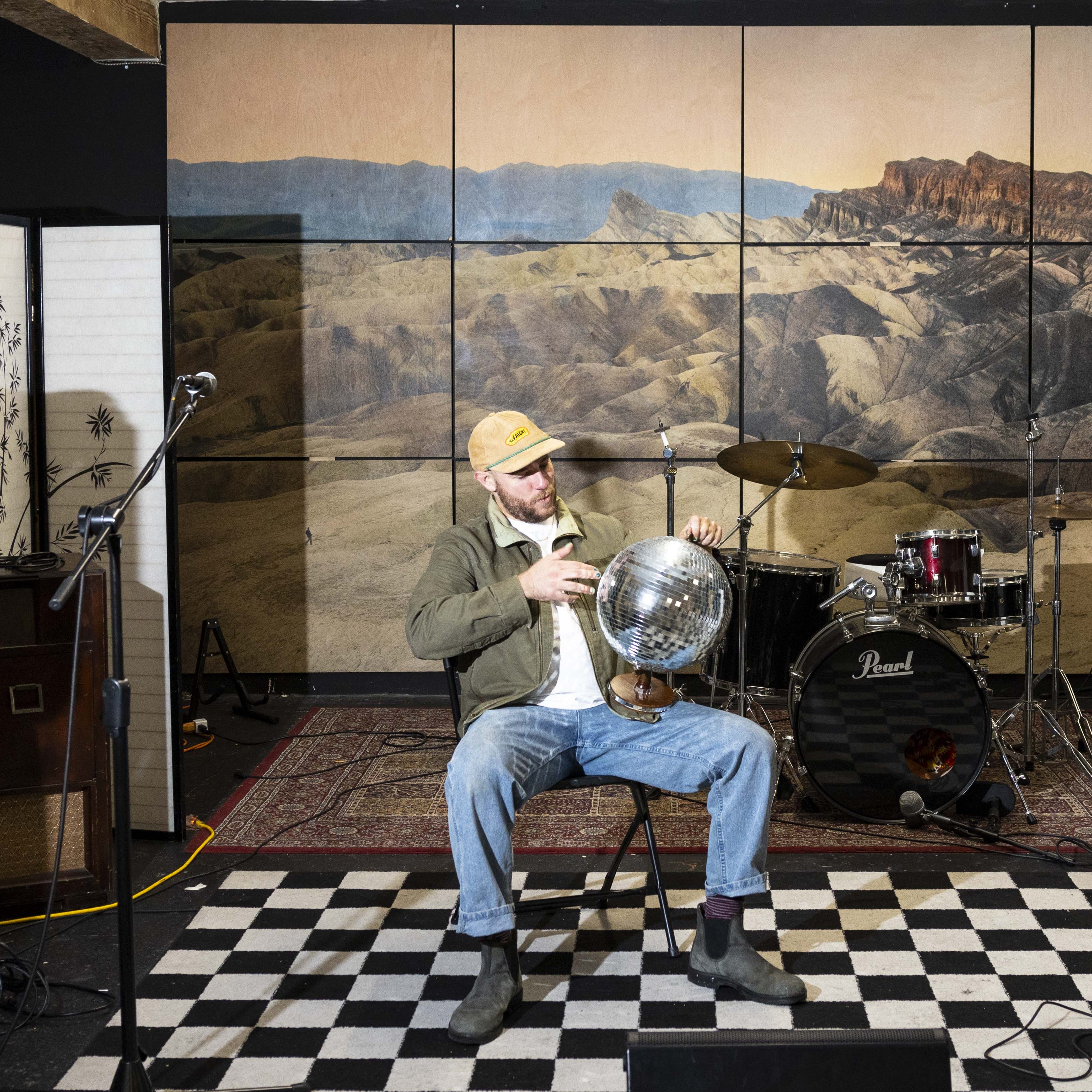 A man sits on a checkered floor, holding a disco ball in a rehearsal room with a desert mural, drums, and microphones around him.