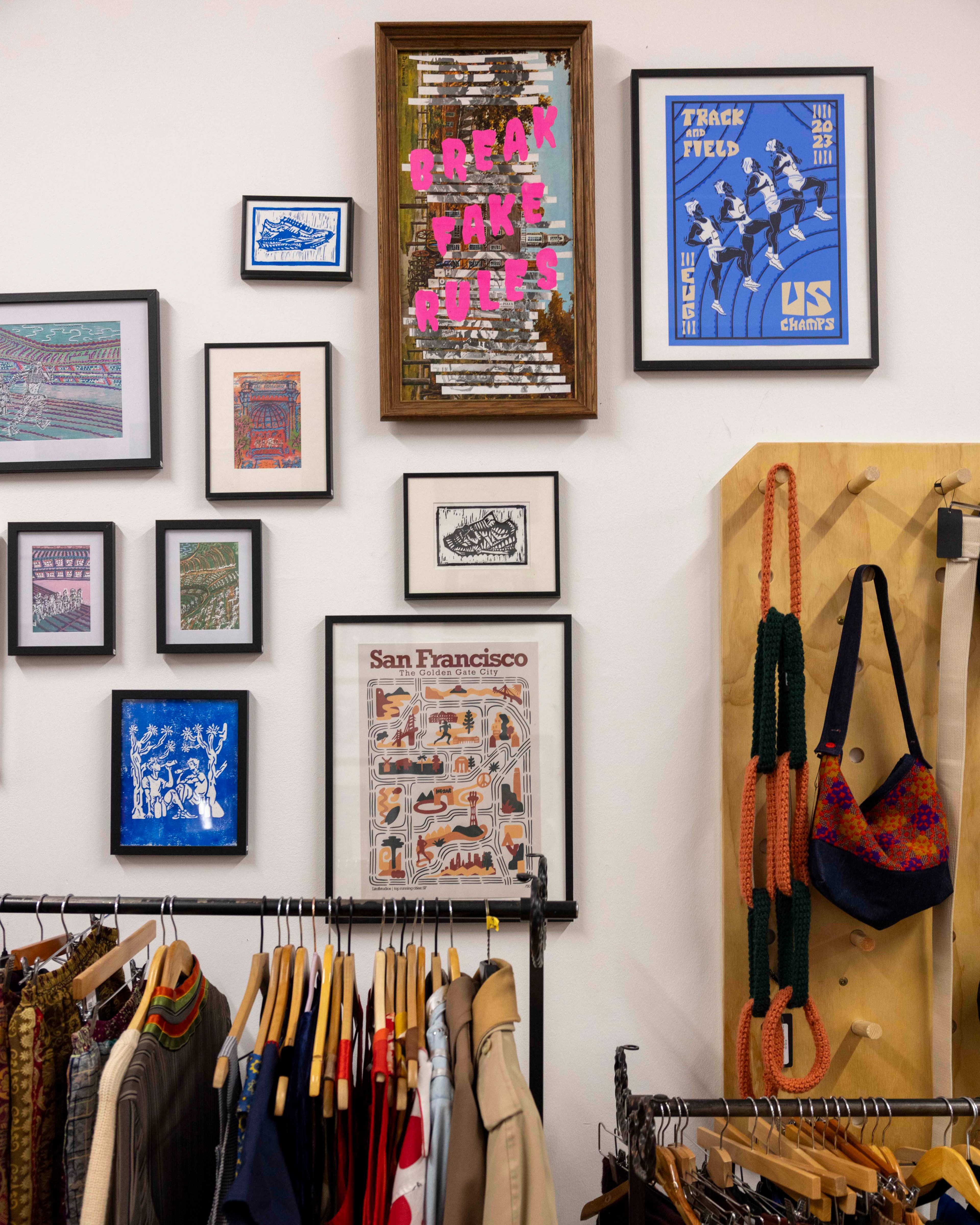 A wall with framed art and prints, including one saying &quot;Break Fake Rules.&quot; Below, a rack holds colorful clothes, and to the right, a purse hangs on pegs.