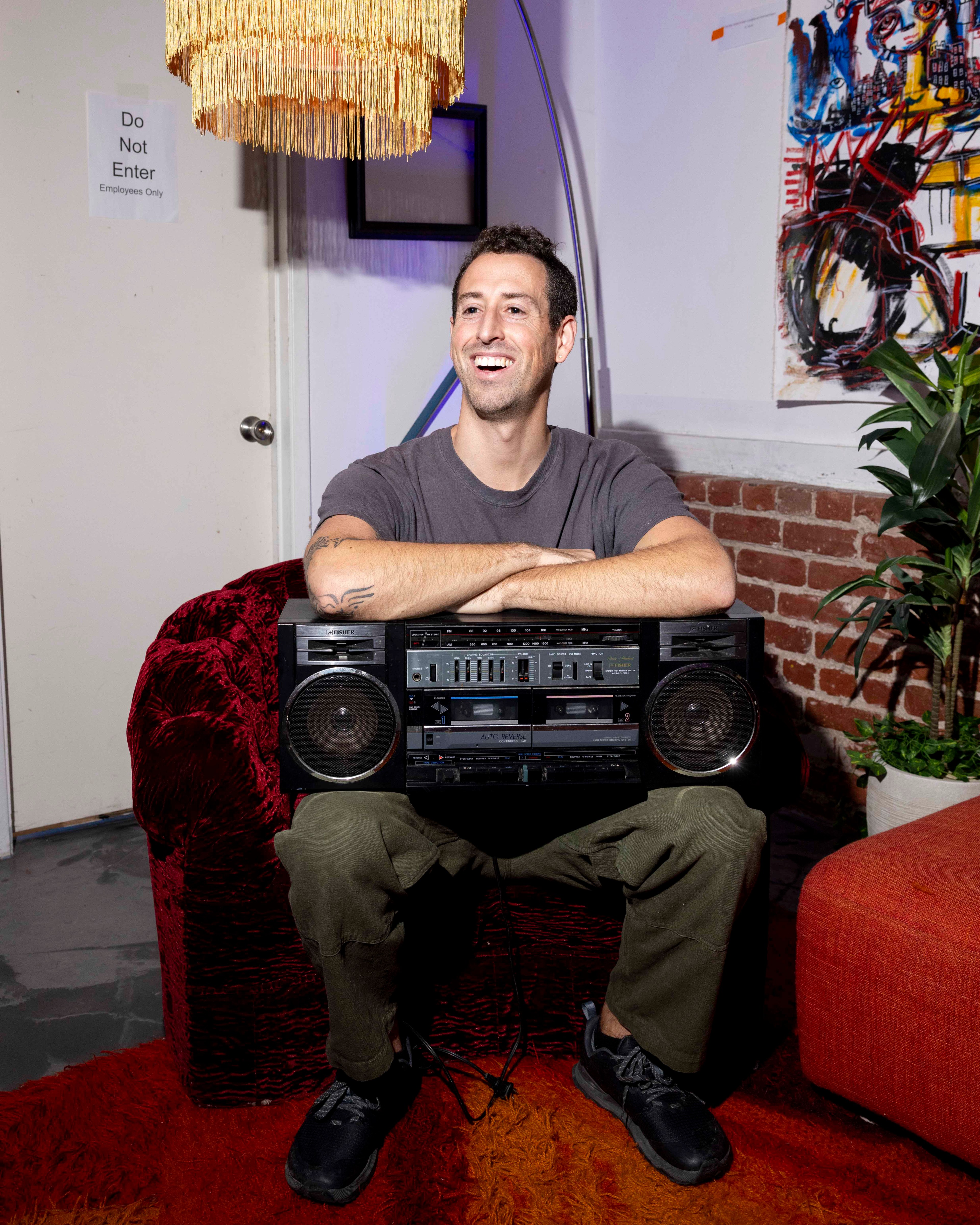 A man smiles while sitting on a red chair, holding a large boombox. There's a fringed lamp, a &quot;Do Not Enter&quot; sign, abstract wall art, and a potted plant nearby.