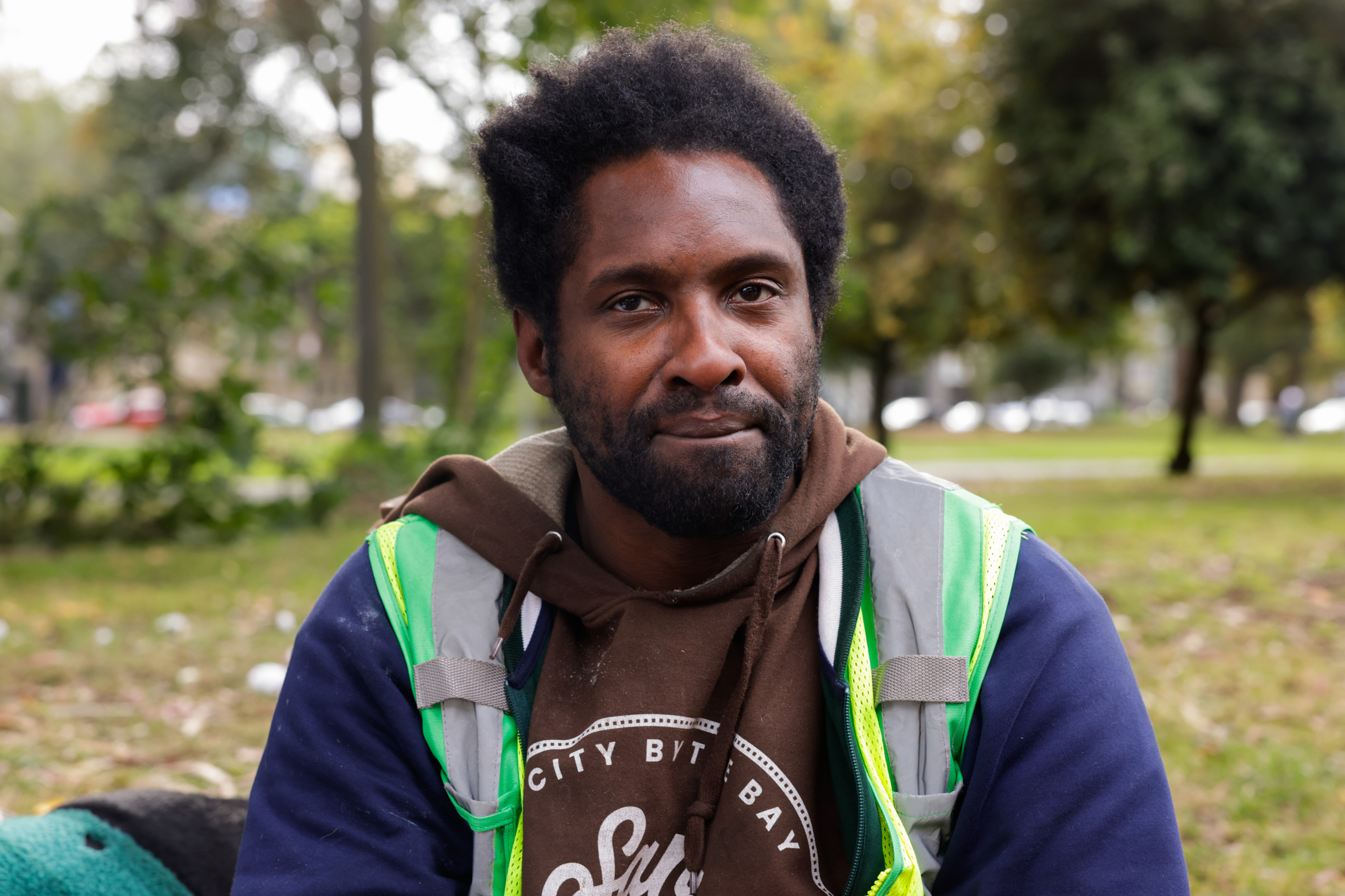 A person with short curly hair wearing a brown hoodie and a reflective vest sits outdoors in a park setting, with trees and grass in the background.