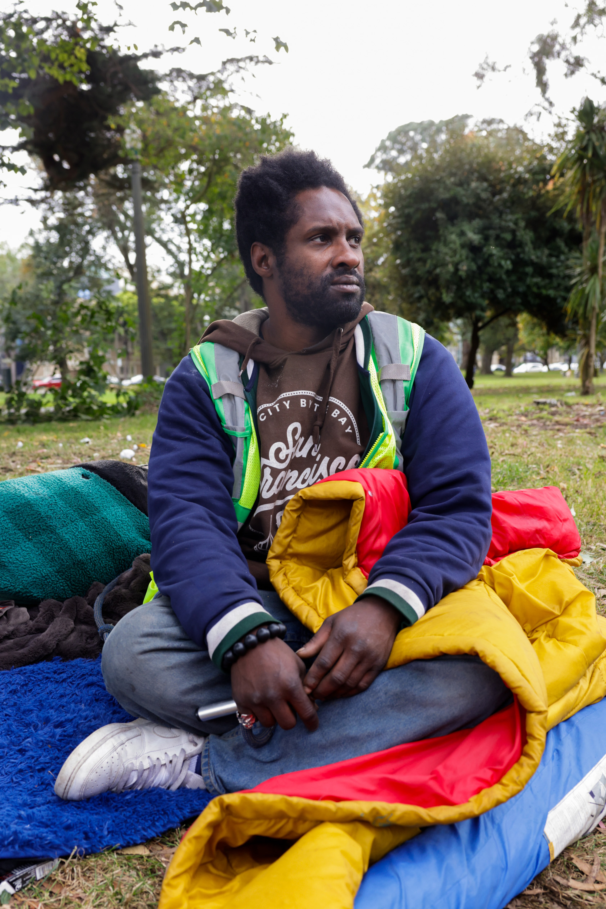 A person sits on the grass, wrapped in a colorful sleeping bag, wearing a jacket and holding a cylindrical object. Trees and greenery surround them in the background.