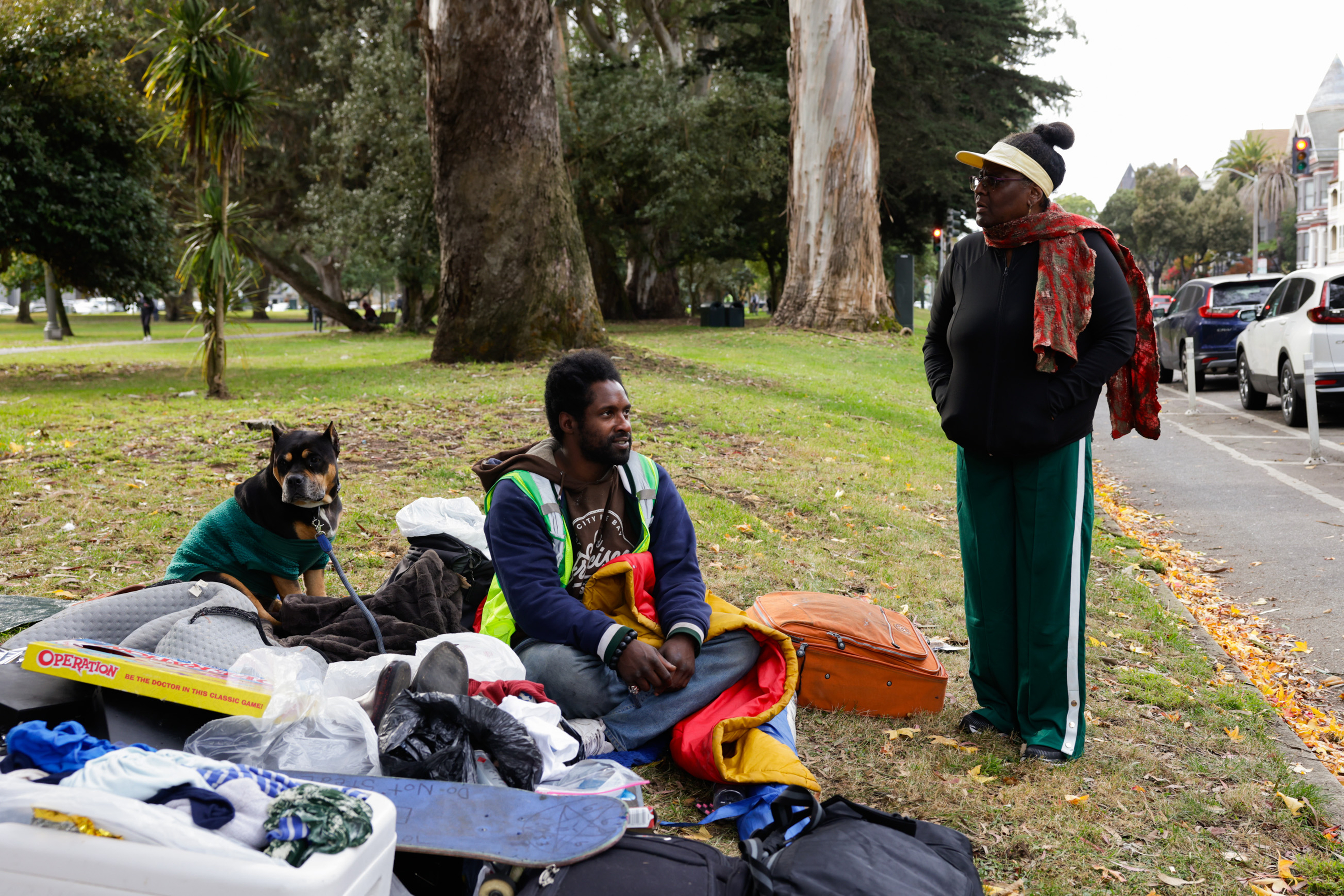 A man sits on the grass with belongings, including a skateboard and a suitcase. A dog in a sweater sits nearby, and a woman stands, facing him in conversation.