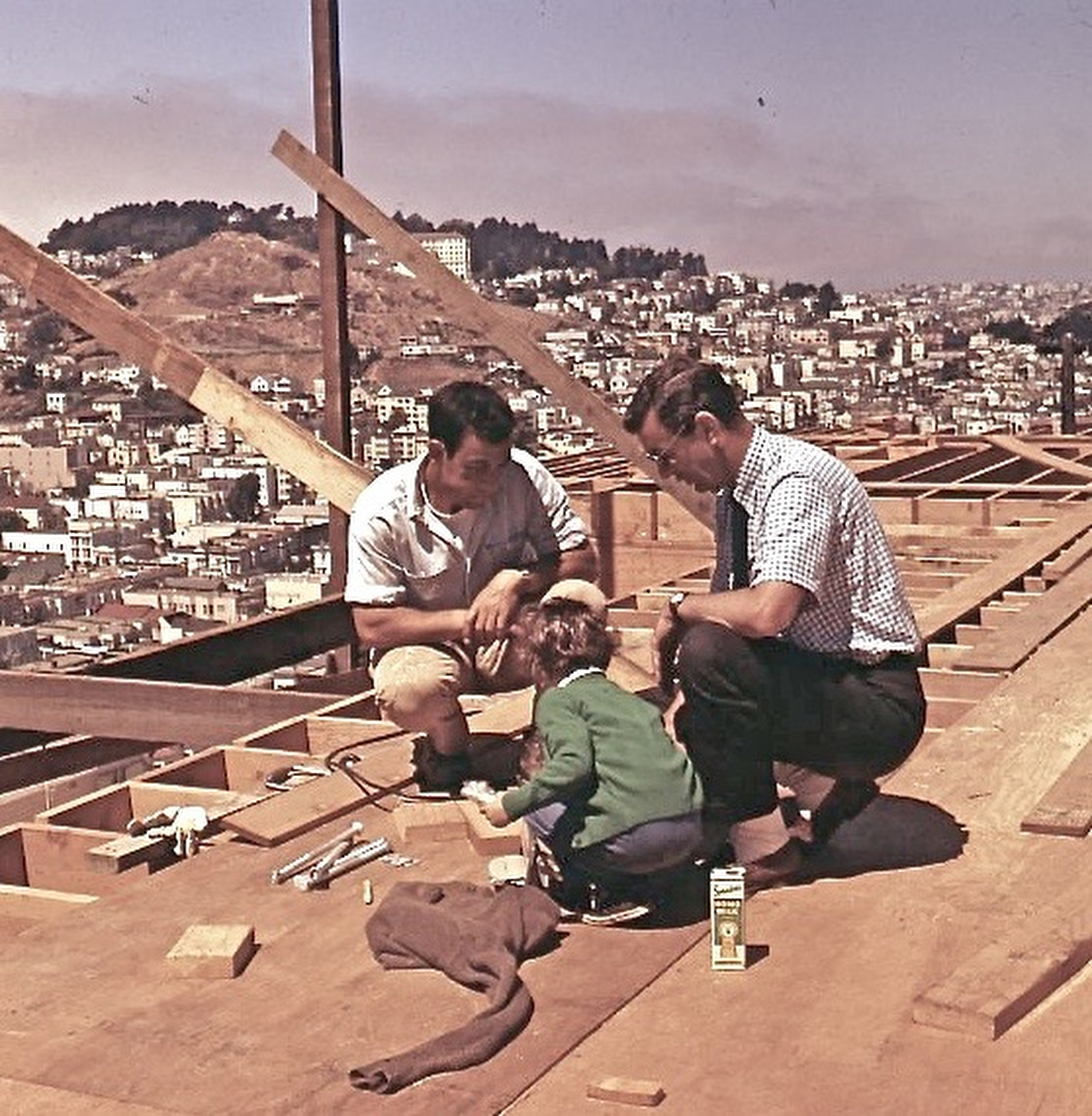 A child and two men are on a wooden construction site, with a view of a hilly cityscape in the background. They're surrounded by tools and a juice box.