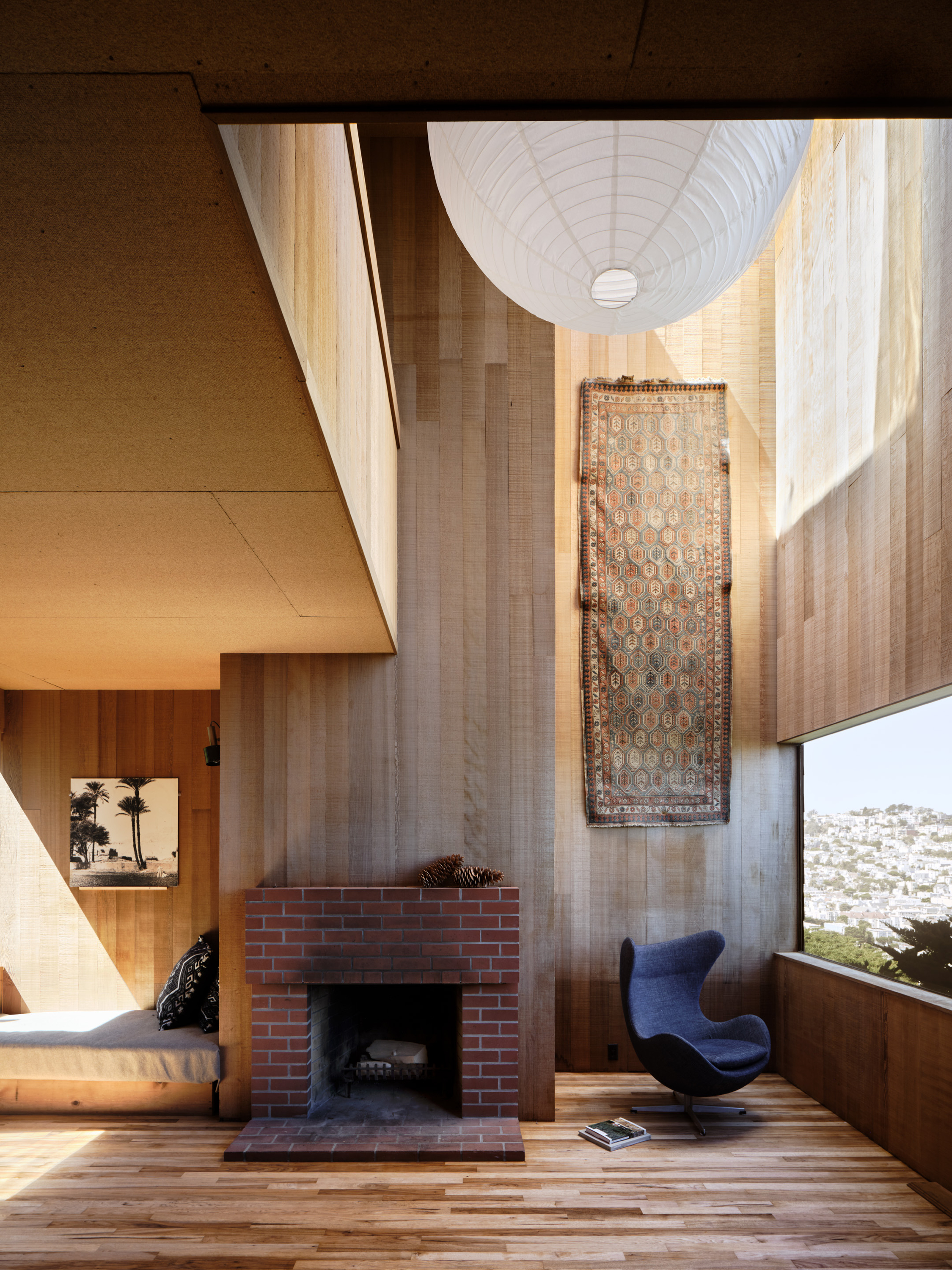 A modern room with wood paneling features a brick fireplace, a cozy corner with a bench, a blue armchair, and a large window with a city view.