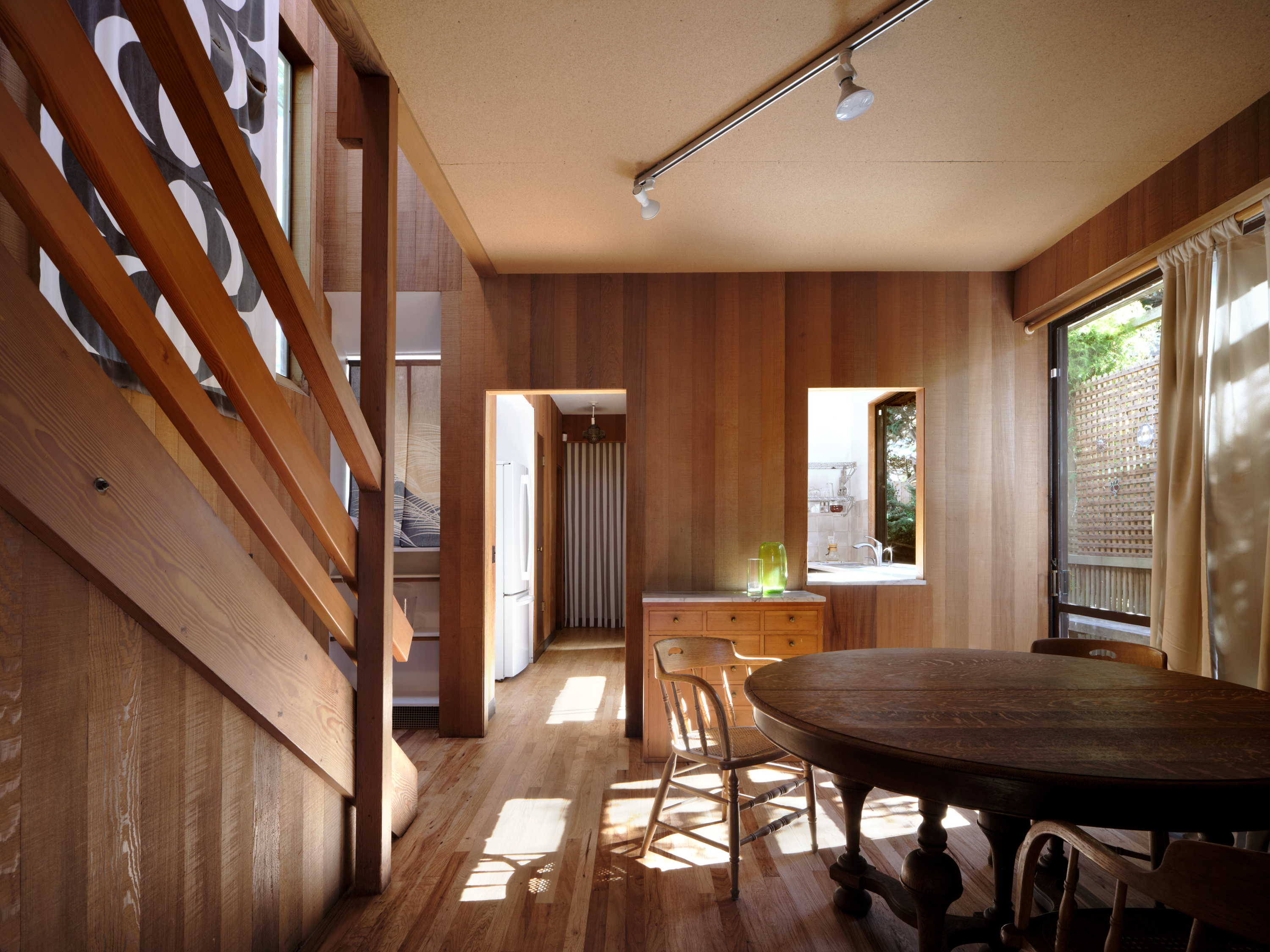 This image shows a cozy wooden interior with a round dining table, chairs, and a staircase. Natural light streams through a window, highlighting warm tones.