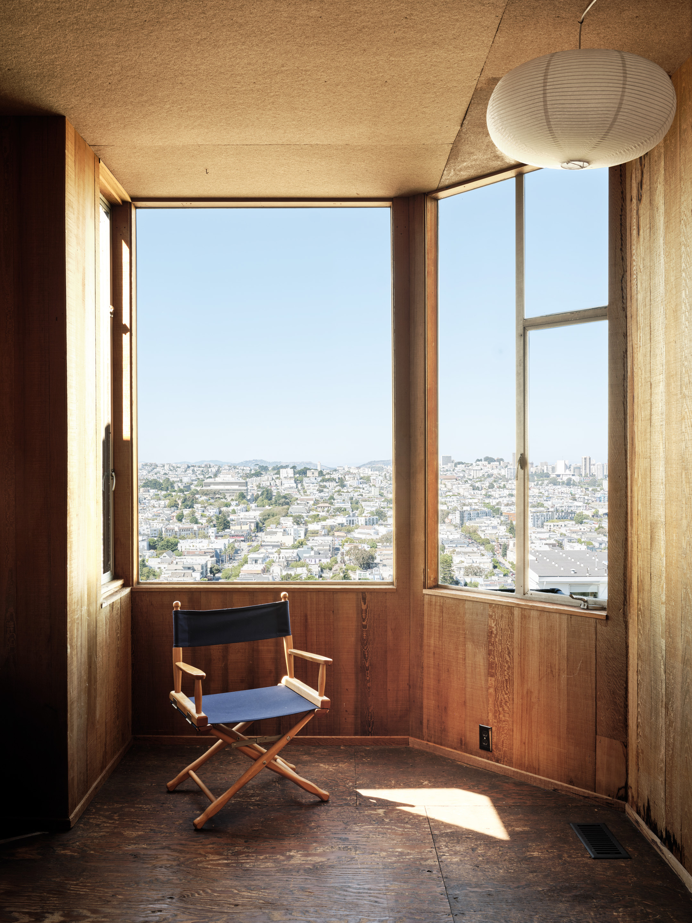 A cozy room features wood paneling and a large window offering a city view. A blue director's chair sits by the window, and a paper lantern hangs from the ceiling.