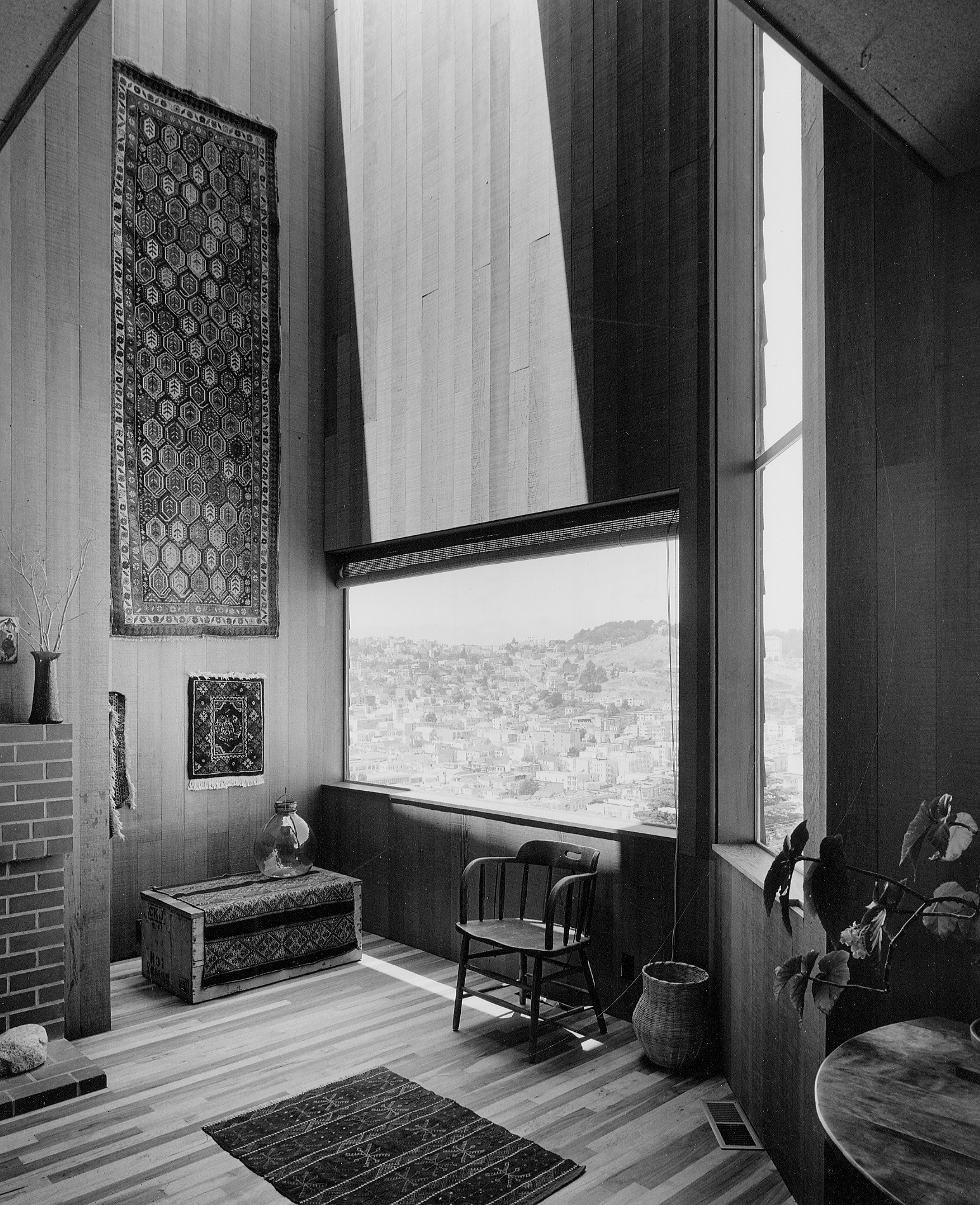 A room with wooden walls features a large window overlooking a city. It includes a wooden chair, woven baskets, decorative rugs, and a brick fireplace.