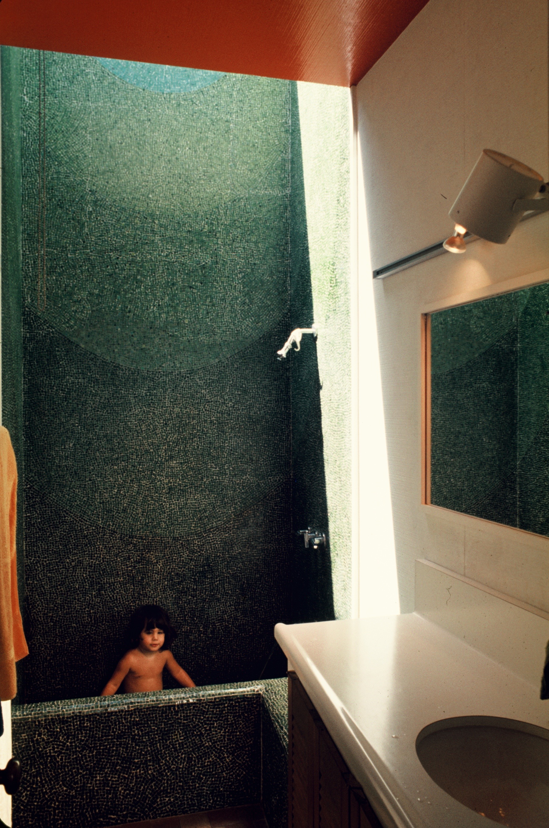 A child is bathing in a green-tiled bathtub under natural light coming from above. The bathroom has a sink with a white countertop and overhead lighting.