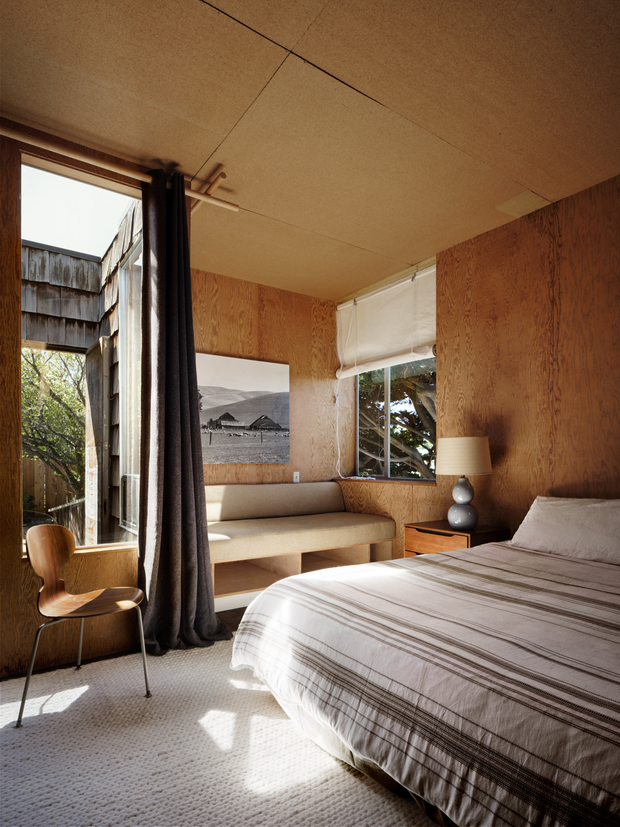 The room features wood-paneled walls, a bed with a striped duvet, a beige cushioned bench, a wooden chair, a lamp, and a large window with sheer curtains.