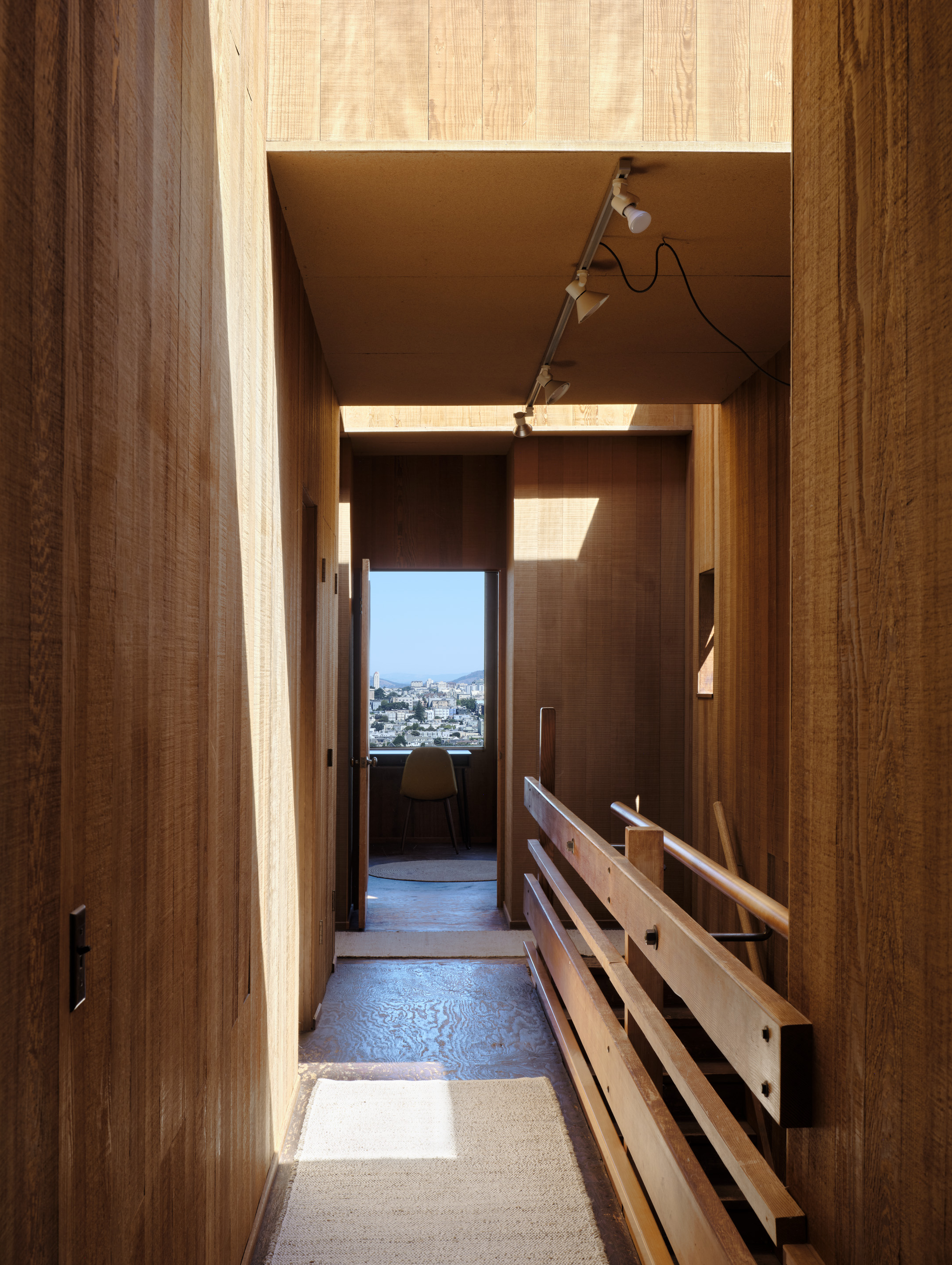 A narrow, wooden hallway with sunlight streaming, leading to a small room with a window view of a cityscape. A simple chair sits by the window.