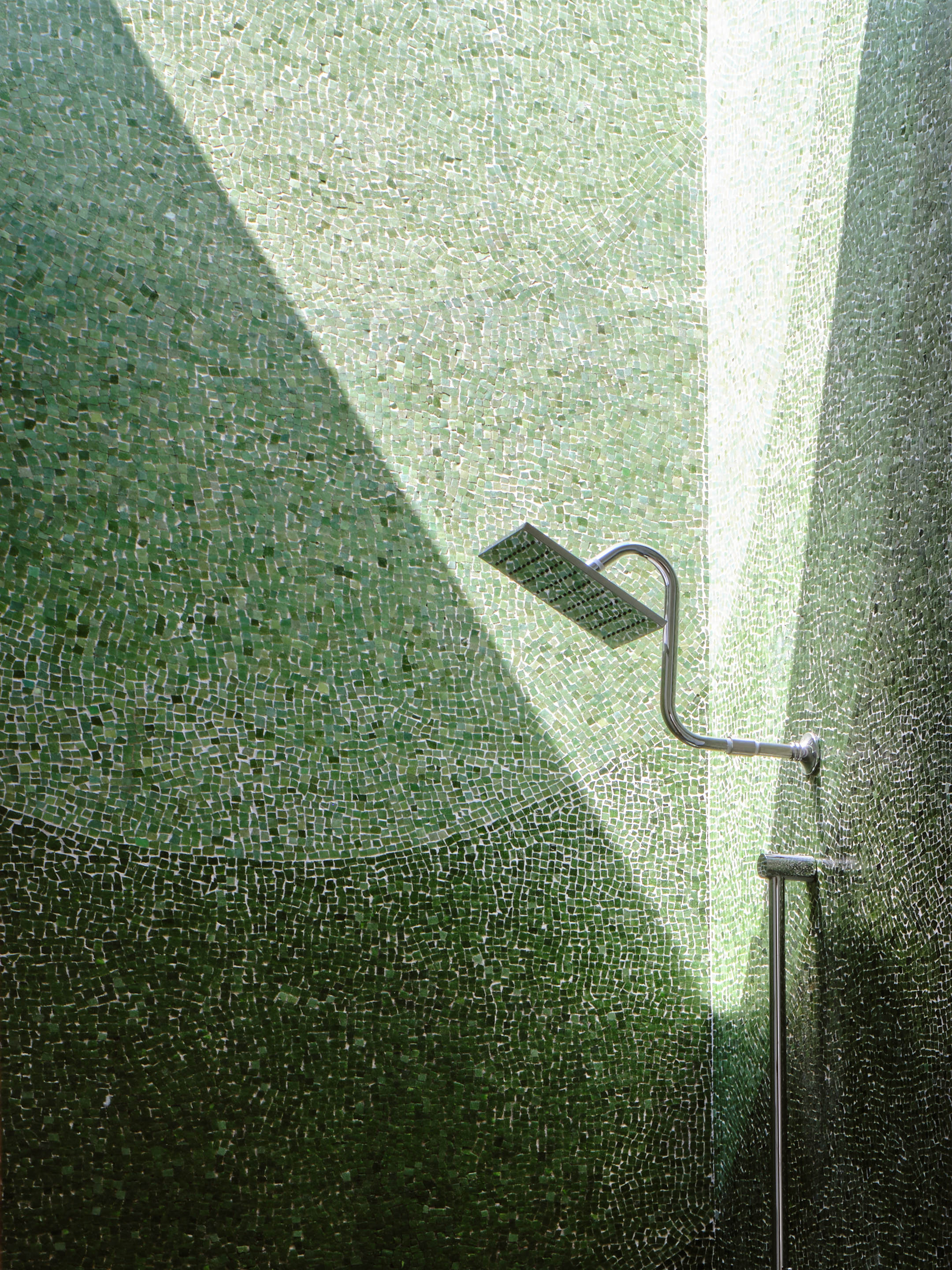 A shower with a square head is set against a wall of small green mosaic tiles. Sunlight creates a bright diagonal band across the tiles.