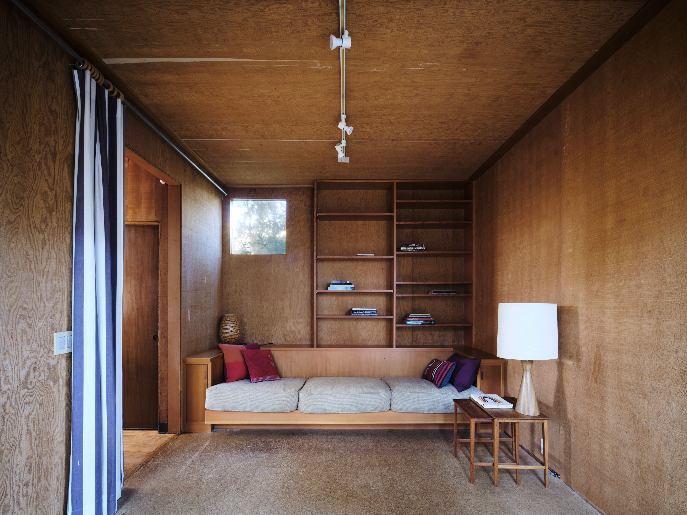 The image shows a cozy room with wood-paneled walls, a beige sofa with colorful cushions, built-in bookshelves, a lamp on a small table, and a striped curtain.