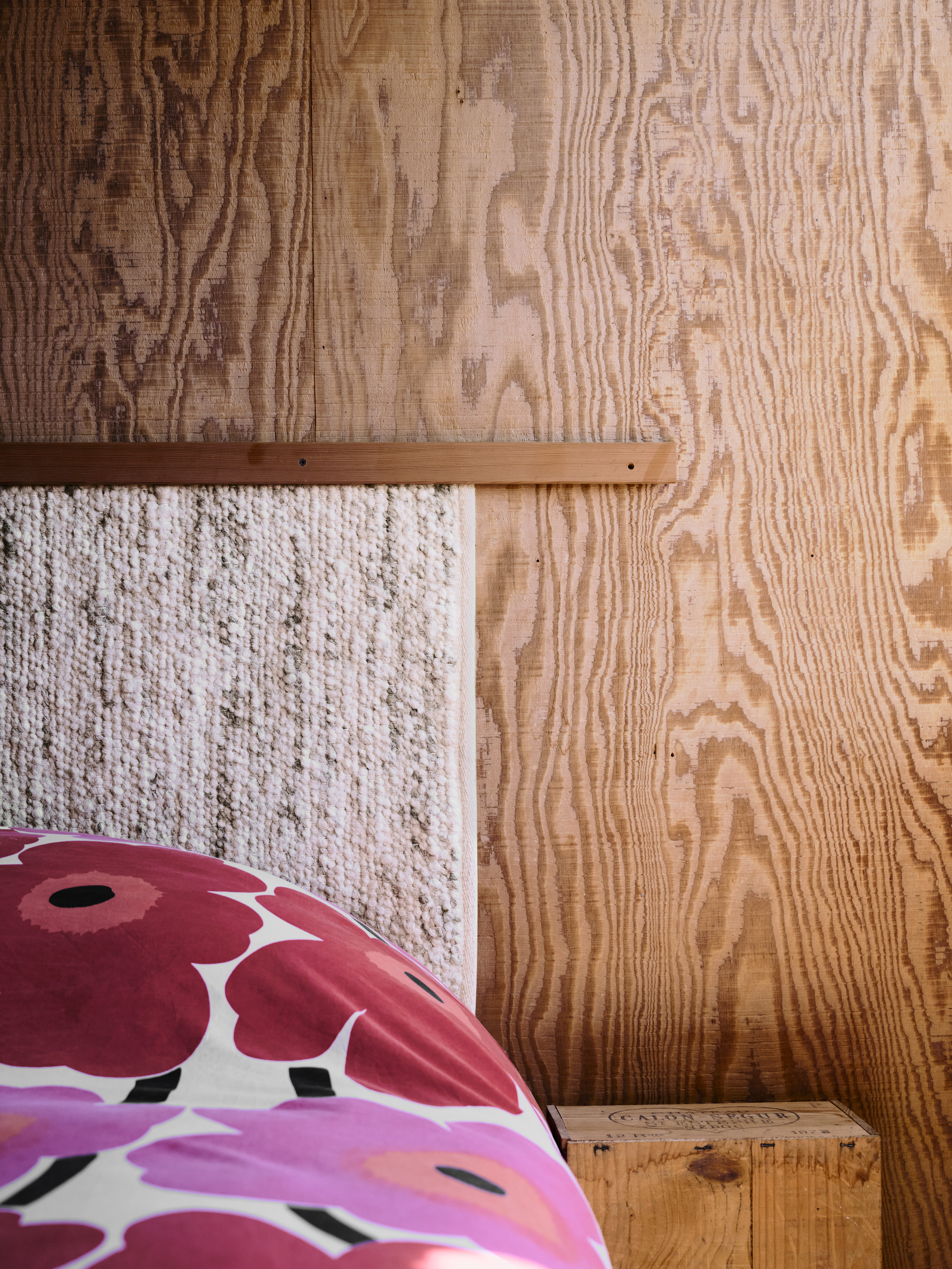 A wooden paneled wall is seen with a textured headboard and a bed adorned with a red floral-patterned blanket. A small wooden crate is next to the bed.