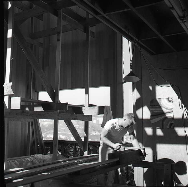 A man is working with wood in a sunlit workshop. Sunlight casts shadows on the walls. There's a construction frame and hanging lamp in the background.