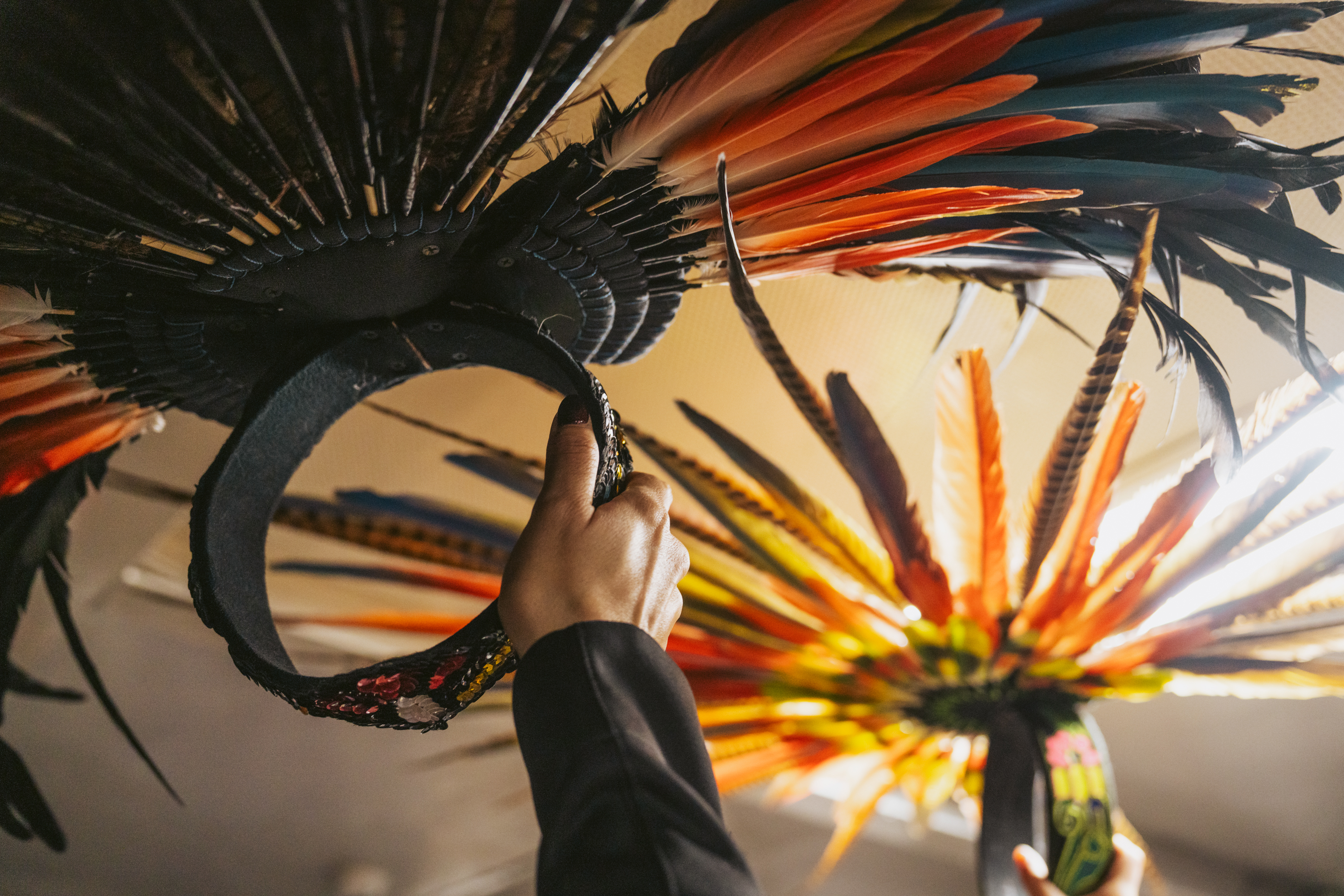 A hand holds an ornate headpiece with vibrant feathers in red, blue, and orange hues against a light background, capturing a cultural or festive essence.