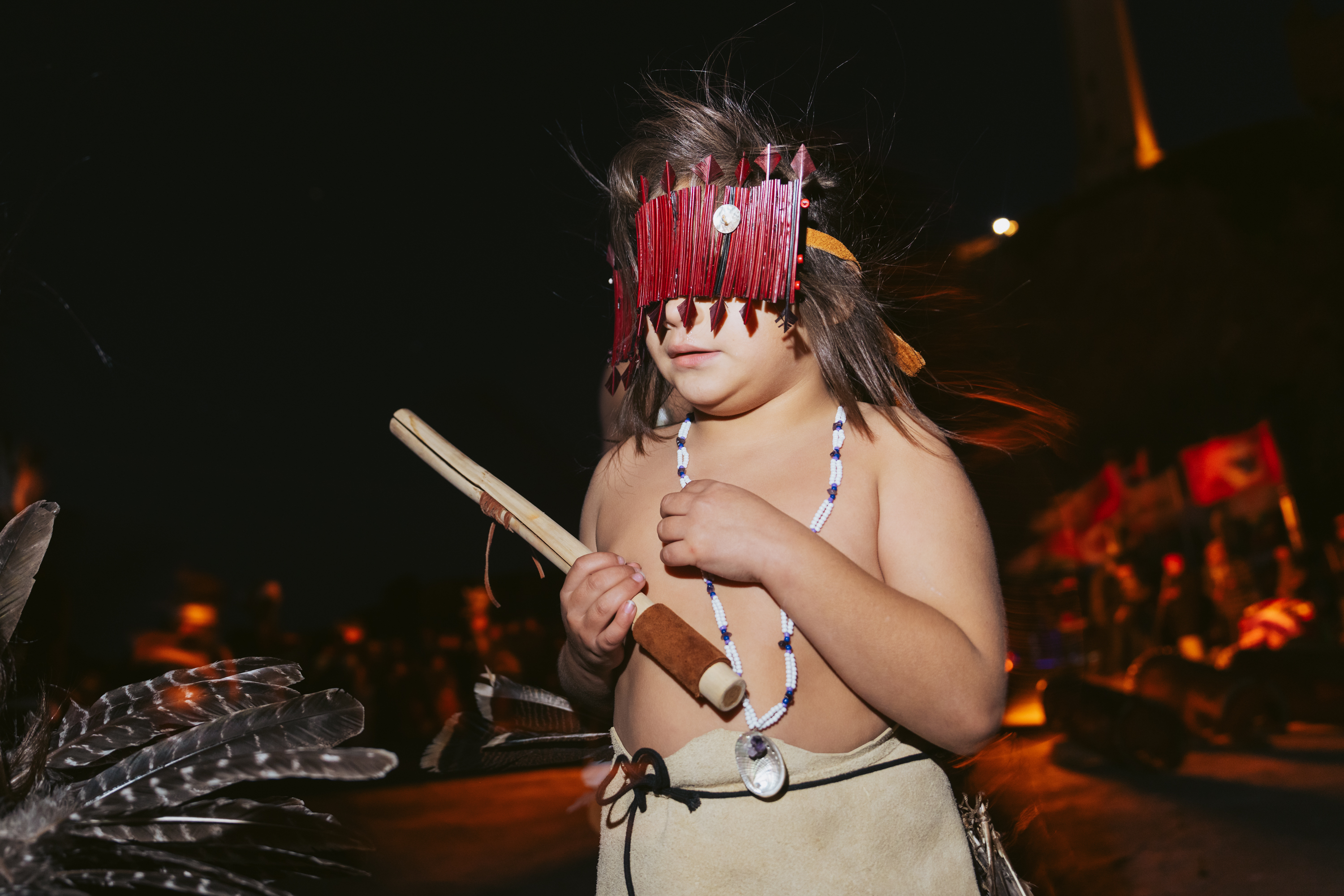 A child wears a traditional red wooden headdress and necklace, holding a stick-like object against a dark background with feathers visible in the foreground.