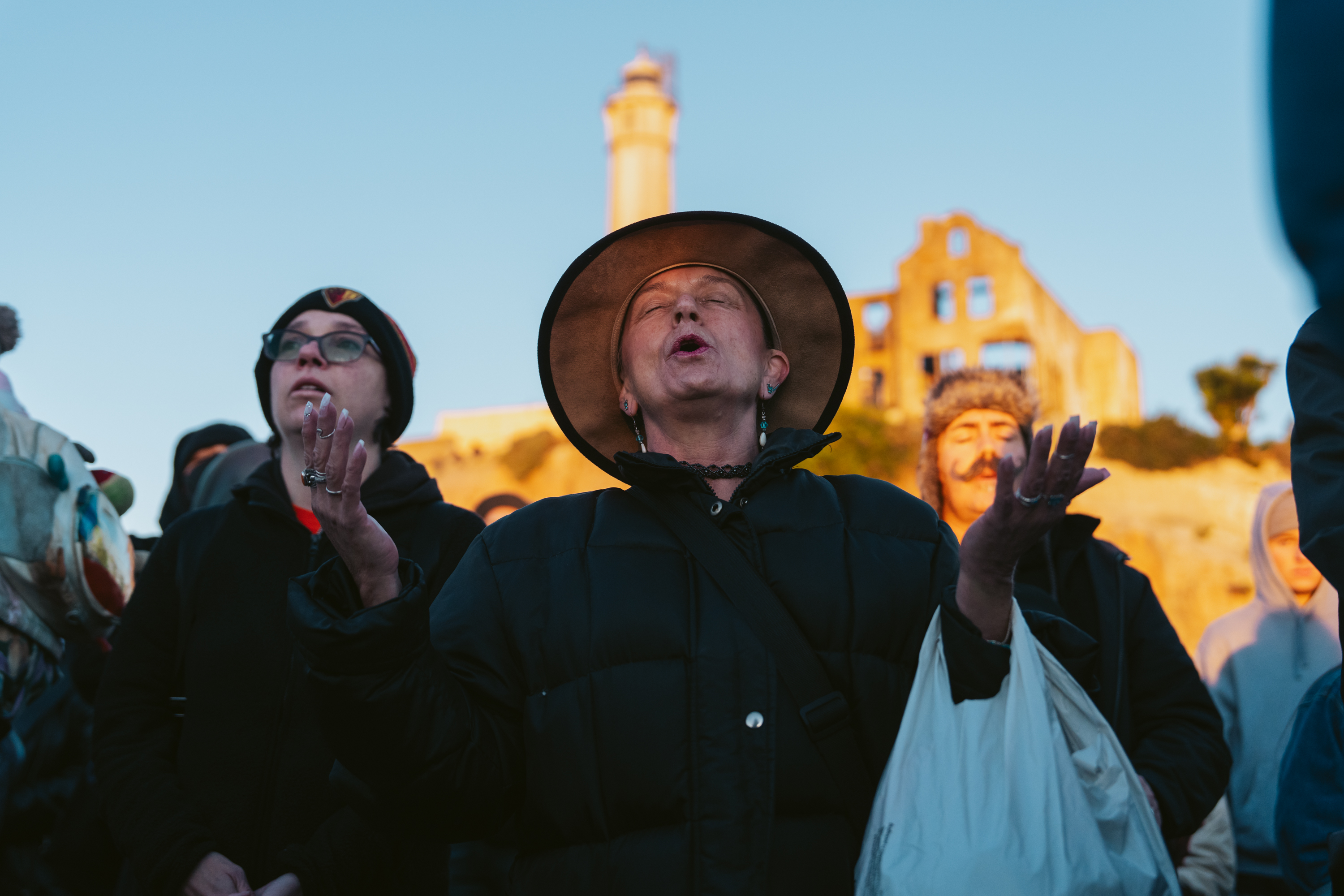 A group of people are outdoors, one with a wide-brimmed hat and eyes closed, raising their hands. A sunny structure and a distant tower are in the background.