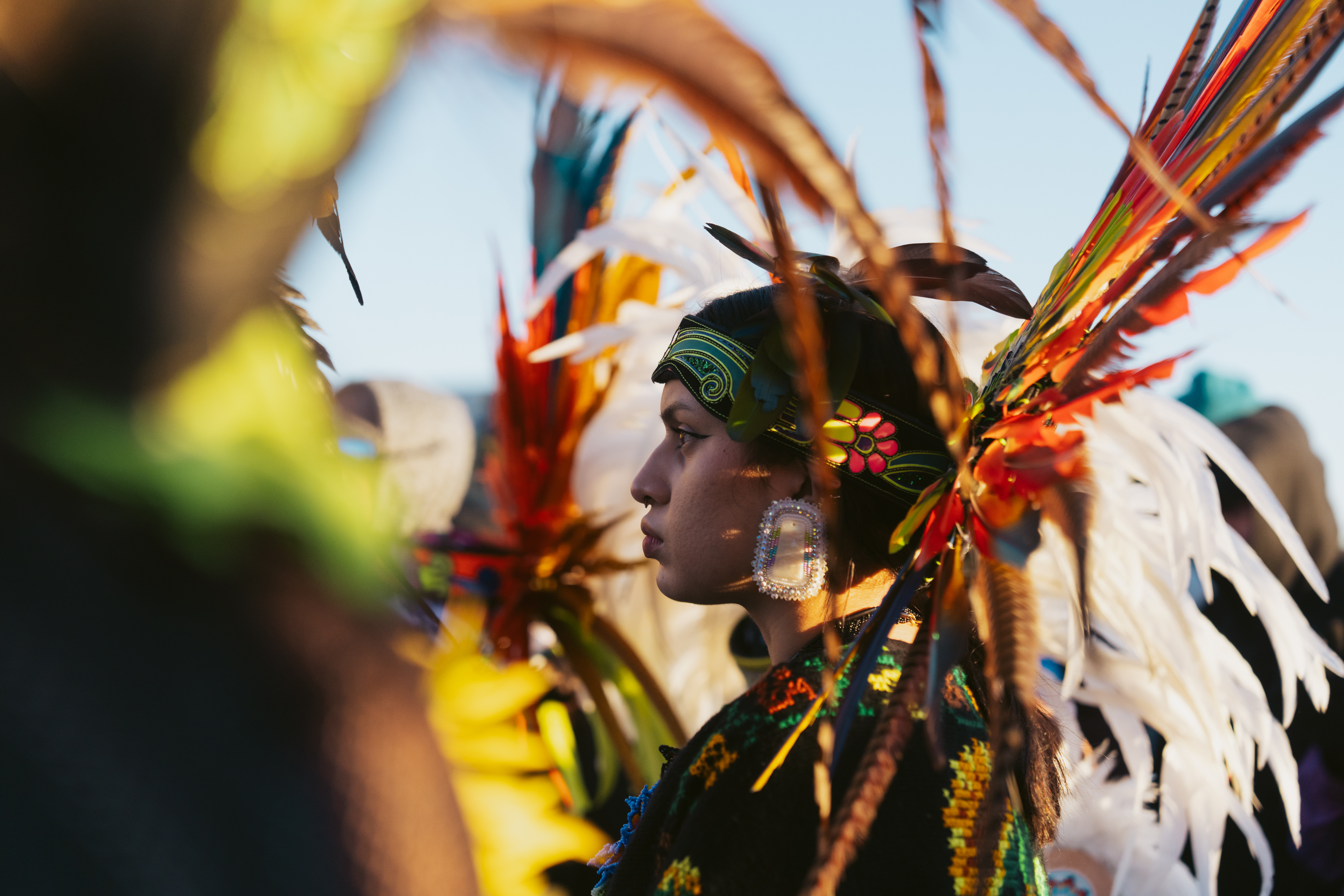 A person wearing a colorful headdress made of feathers and floral designs stands in profile. Their attire features intricate patterns, and large beaded earrings are visible.