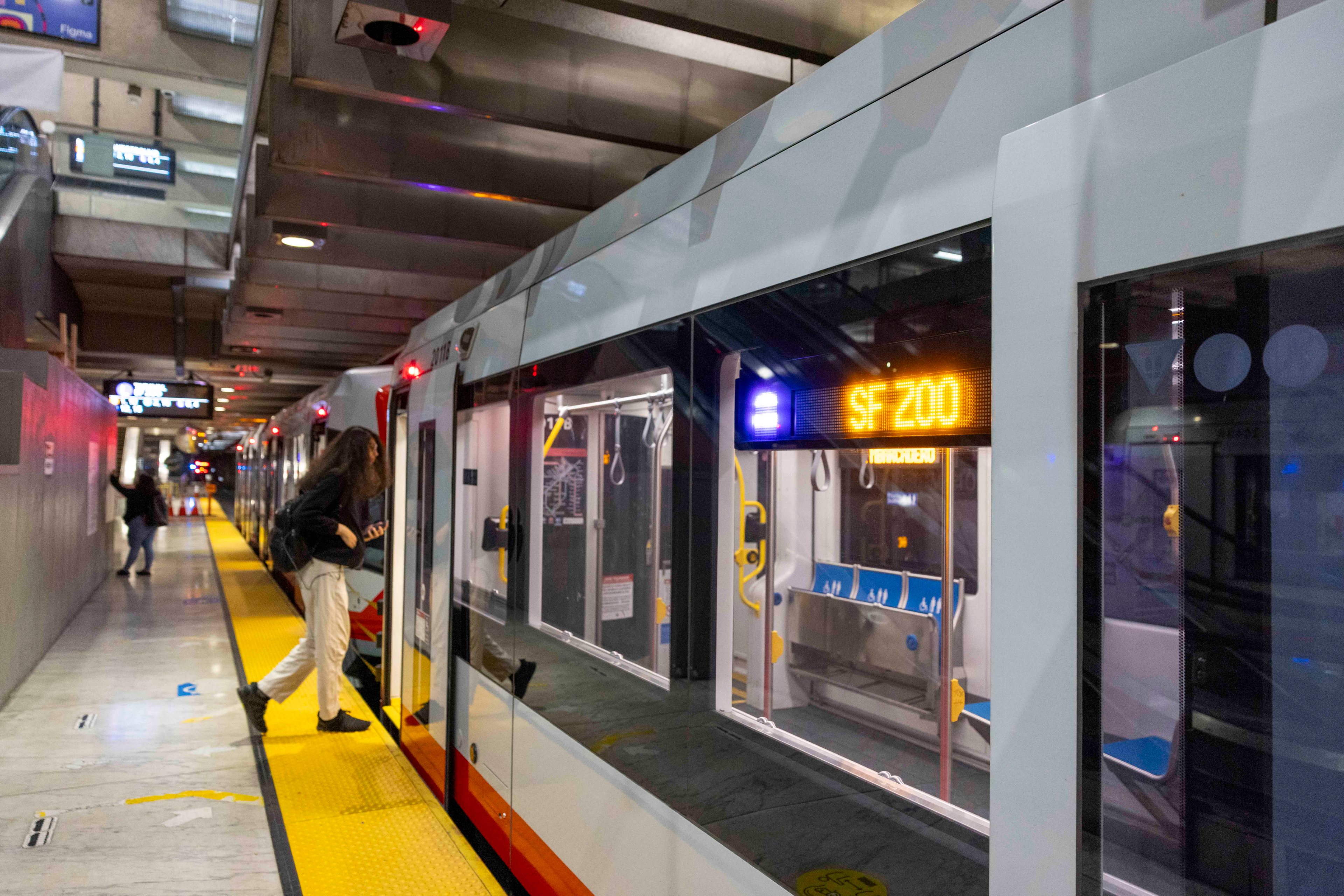 A person boards a modern train at a station. The train's destination sign reads &quot;SF ZOO.&quot; The platform has a yellow safety edge and overhead lights.