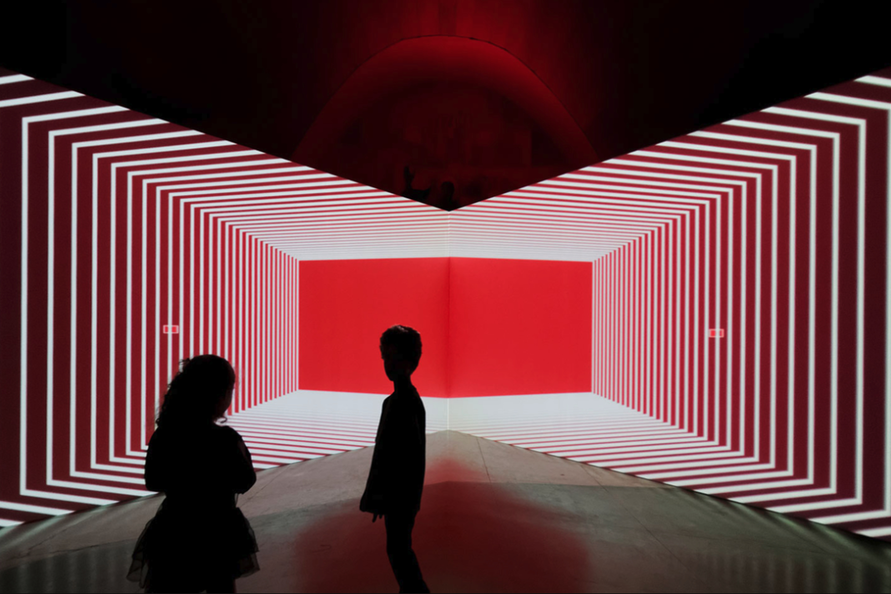 Silhouettes of two people stand before an abstract, immersive art installation featuring a geometric red and white tunnel with concentric lines.