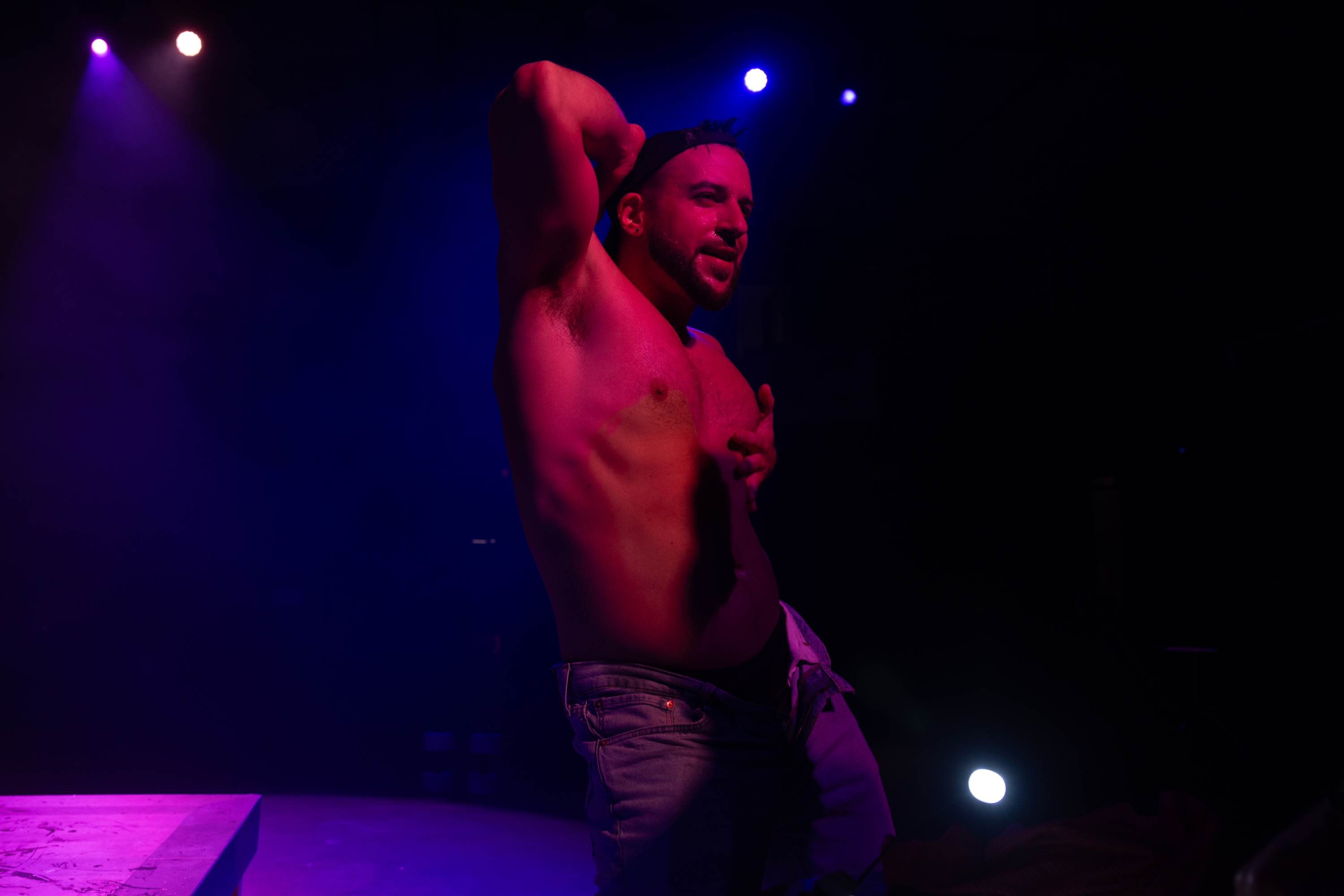 A person, shirtless and in jeans, poses confidently on stage with vibrant purple and blue lights creating a dynamic atmosphere.