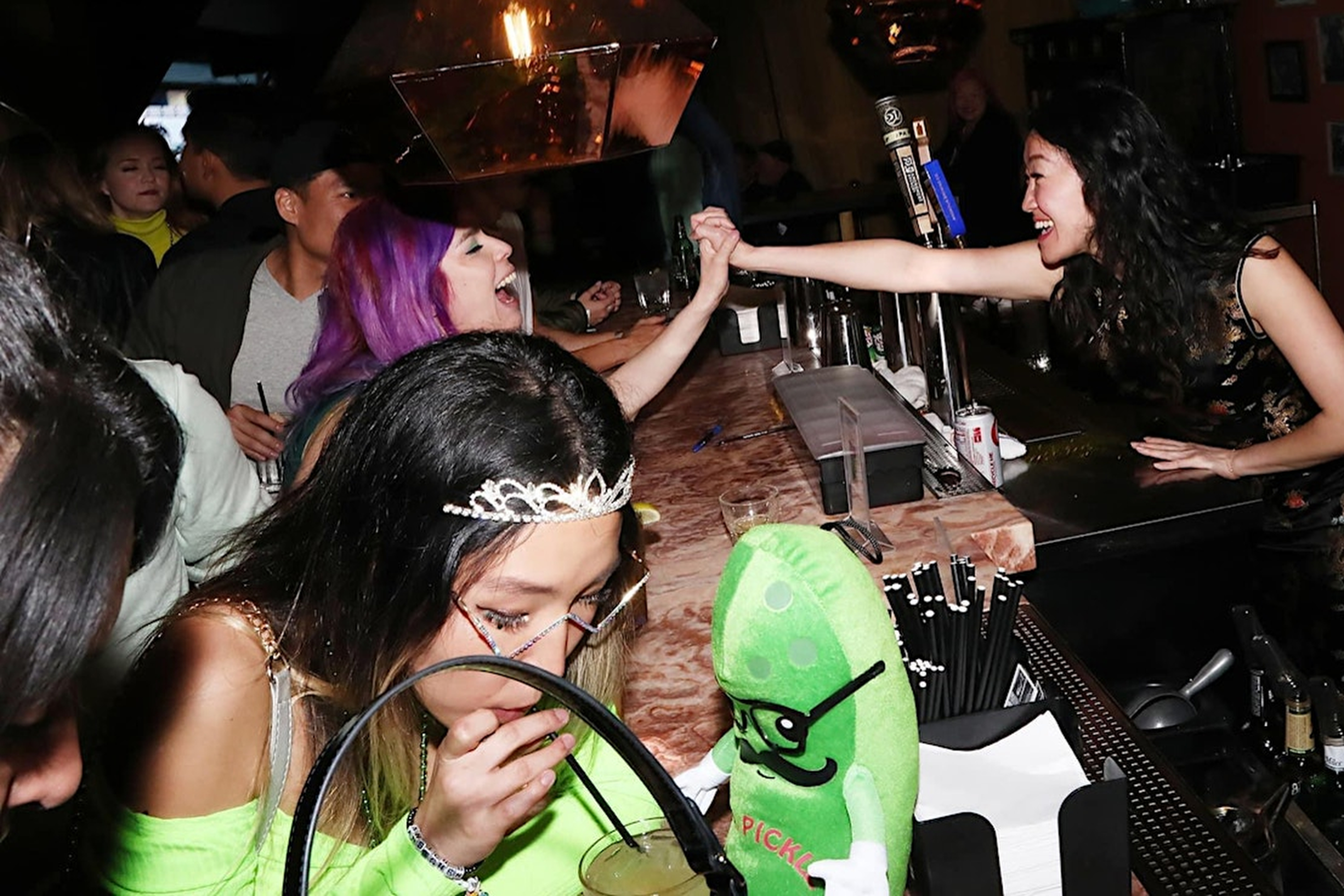 People are having a lively time at a bar. One woman with purple hair is laughing and arm wrestling with a bartender. Others are enjoying drinks, including a woman with a tiara and a stuffed pickle toy nearby.