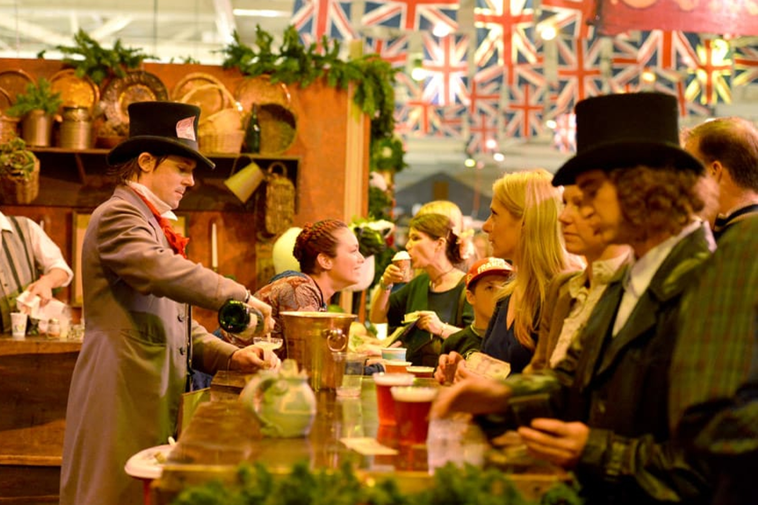 People in Victorian-era costumes gather at a bar with British flags above. A man in a top hat serves drinks while others wait and chat.