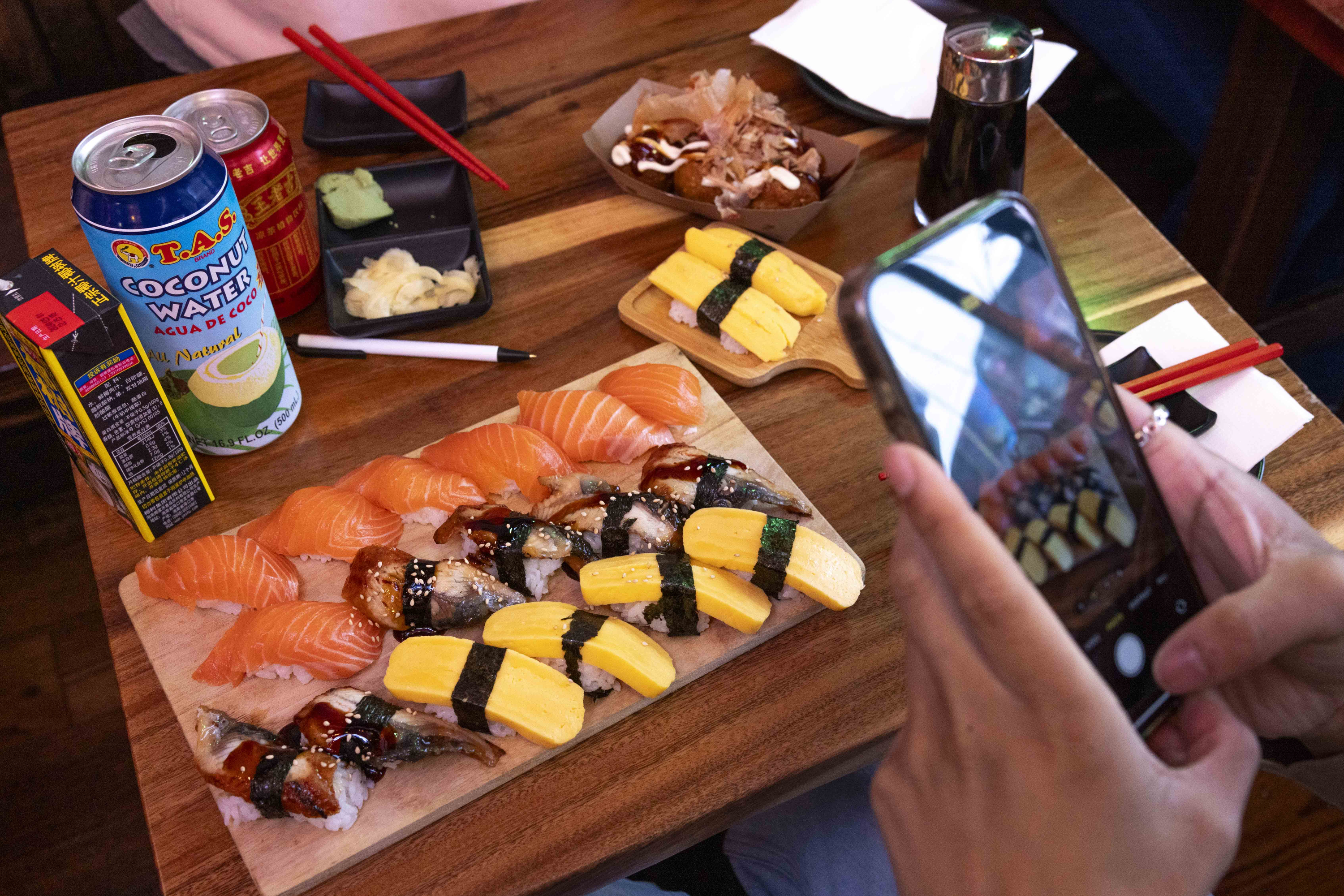 A table with sushi platters, drinks, and condiments. A person is taking a photo of the sushi, which includes salmon, eel, and tamago nigiri.