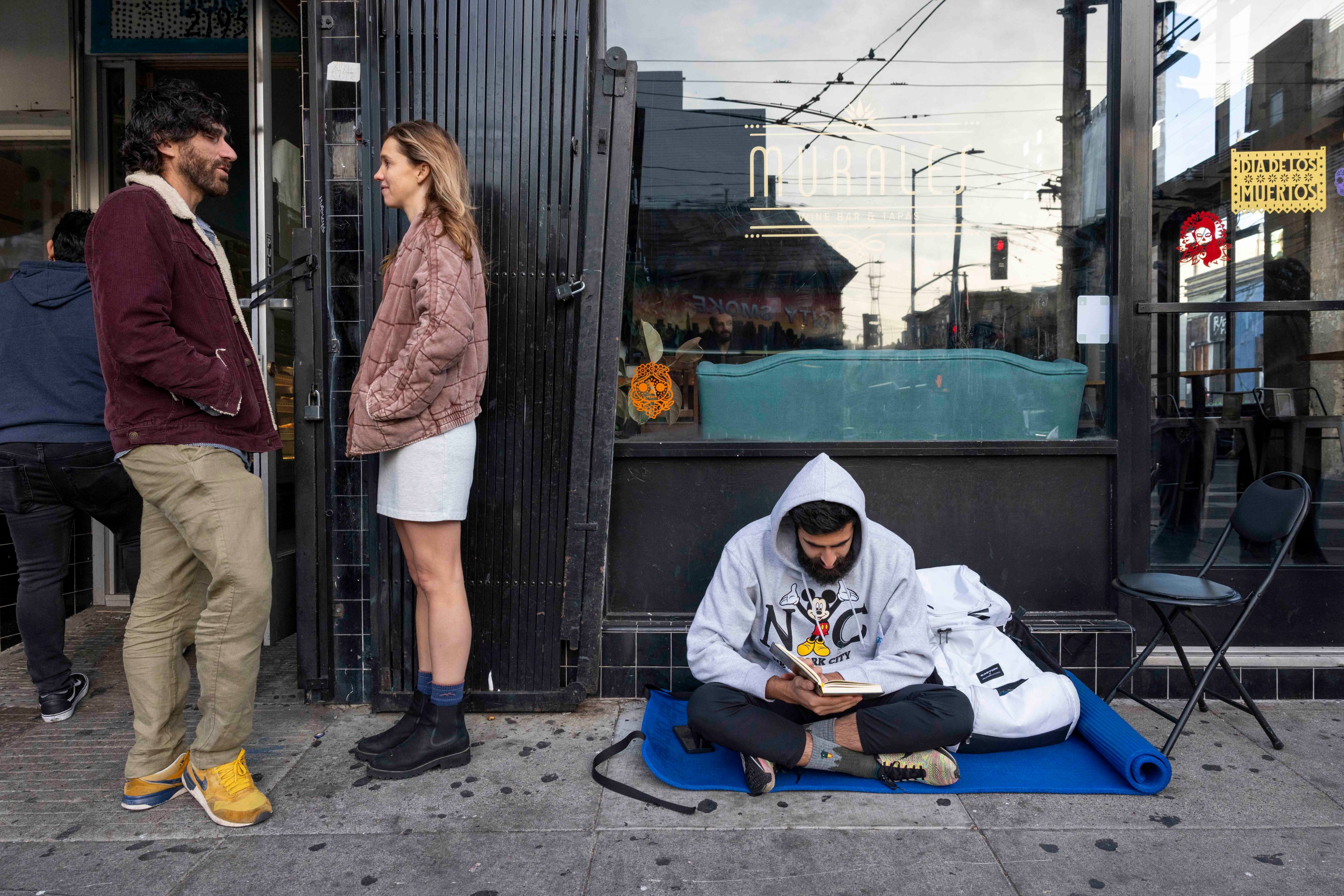 A man and woman stand outside a shop talking, while another man sits on the ground reading a book. He's wearing a hoodie and is surrounded by a mat and bag.