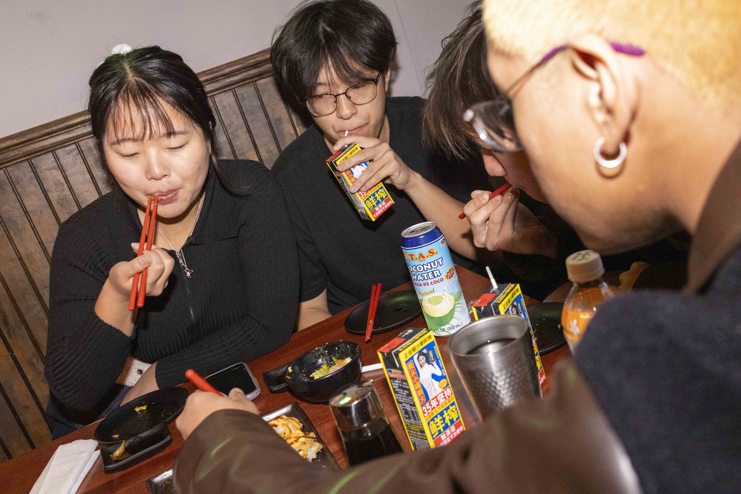 Four people are eating together at a table with snacks, using red chopsticks. They are drinking from cartons and cans, and there are various drinks nearby.