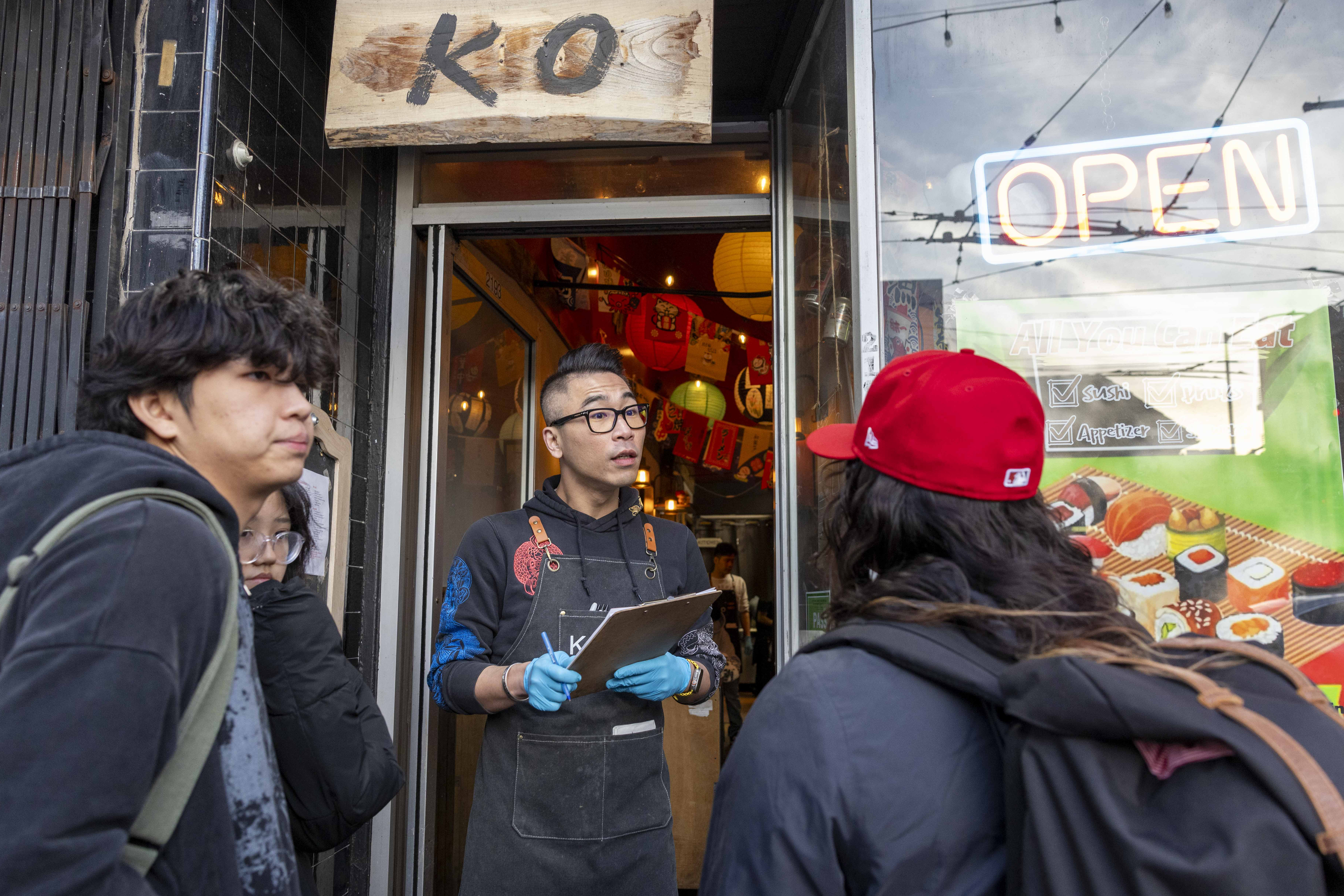 A person wearing gloves and holding a clipboard talks to three people outside a sushi restaurant with a wooden &quot;KO&quot; sign and a neon &quot;OPEN&quot; sign.