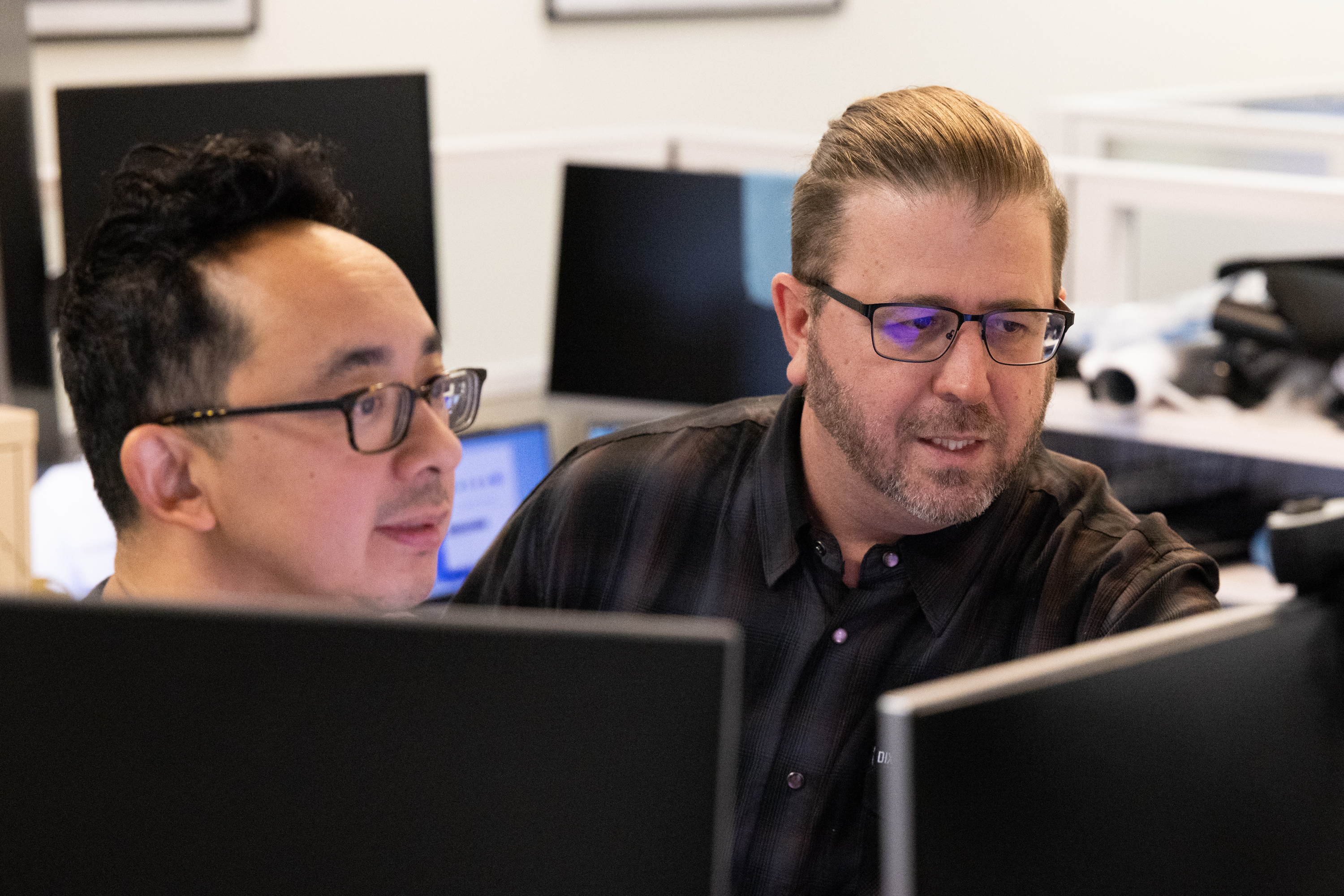 Two men are seated closely together in an office setting, focused on computer screens. They are engaged in a collaborative discussion.