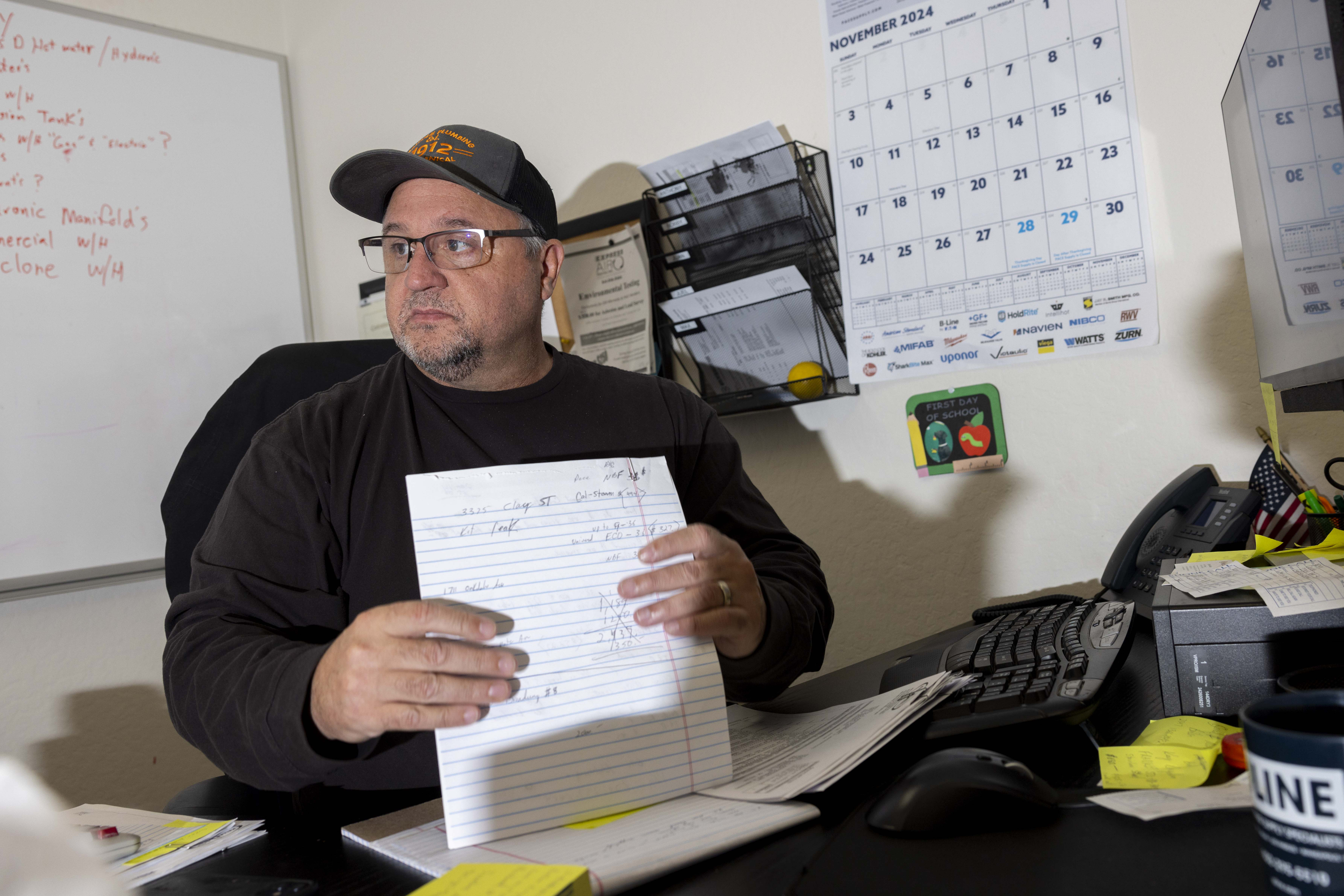 A man wearing glasses and a cap sits at a desk, holding lined paper. Behind him are a calendar and shelves with files, and the desk has papers, a keyboard, and a phone.