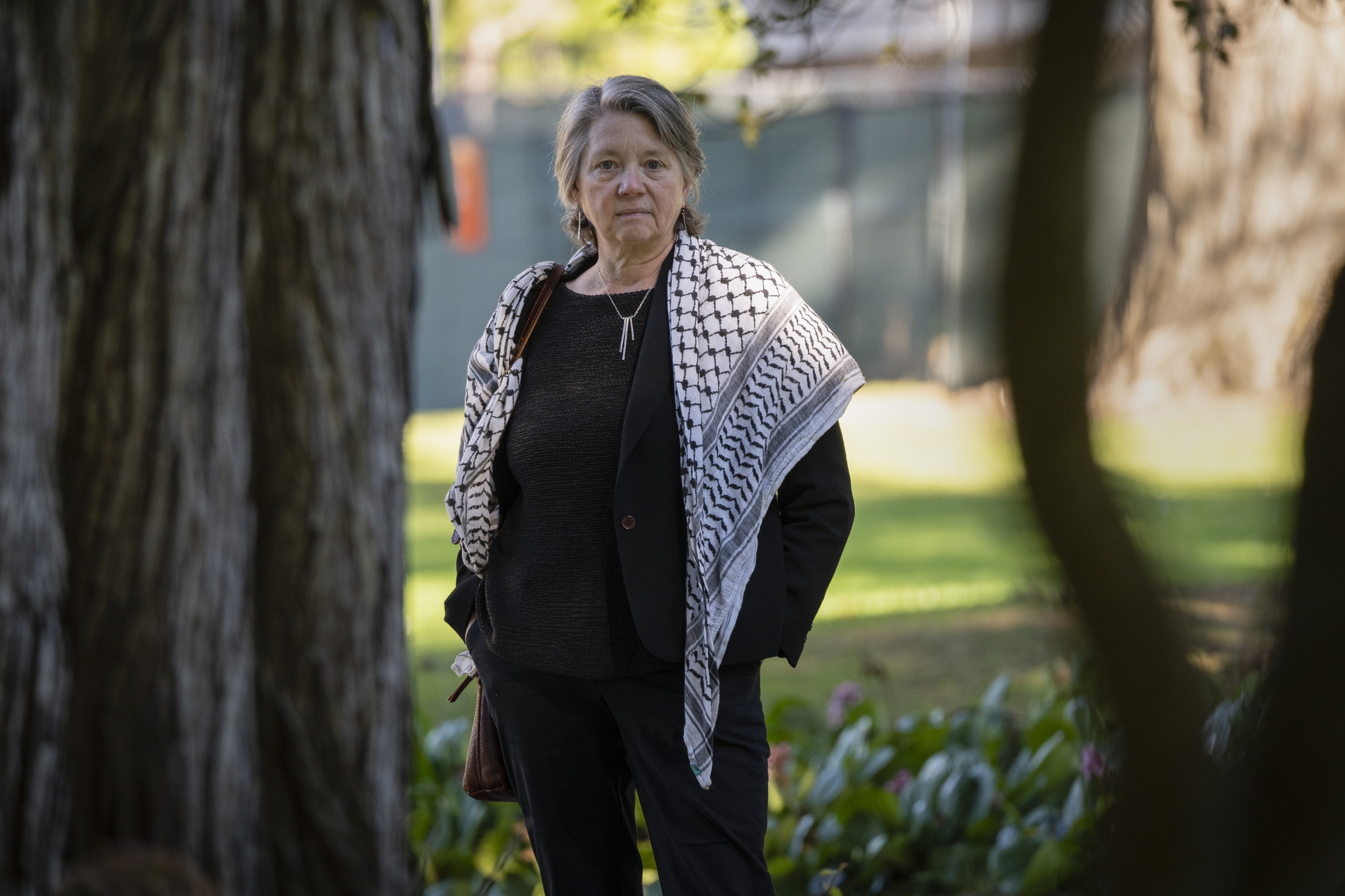 A person stands in a park, wearing a dark outfit and a patterned scarf. They have short, gray hair and the background is filled with trees and greenery.