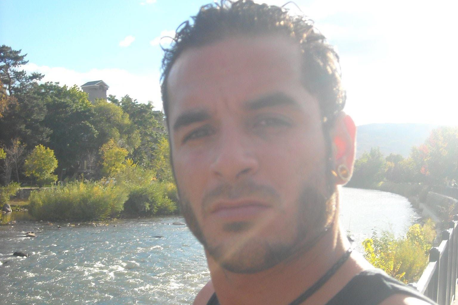 A man with short curly hair and facial hair is taking a selfie by a river on a sunny day. He's wearing a black tank top and necklace; lush greenery surrounds him.