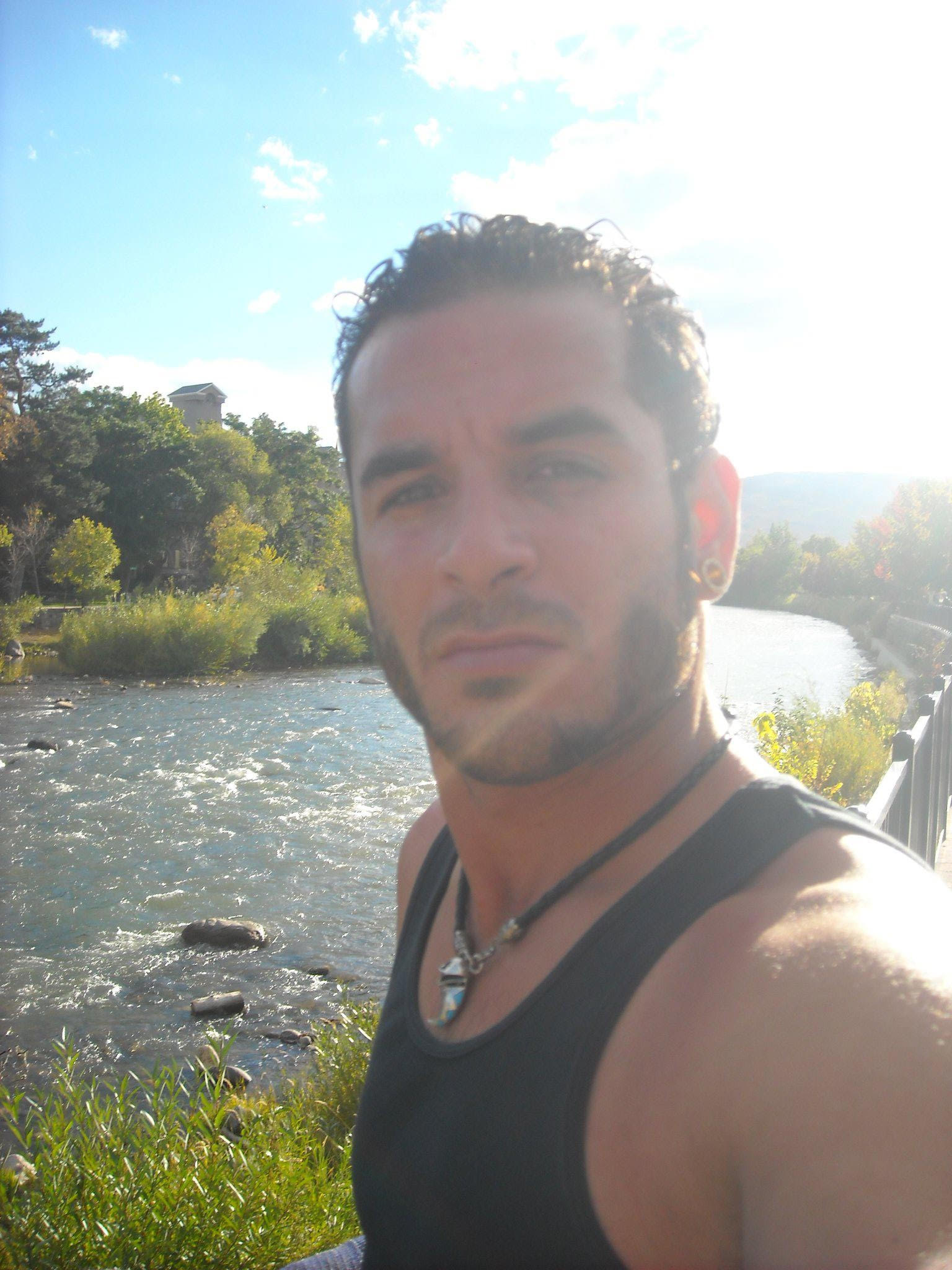 A man with short curly hair and facial hair is taking a selfie by a river on a sunny day. He's wearing a black tank top and necklace; lush greenery surrounds him.