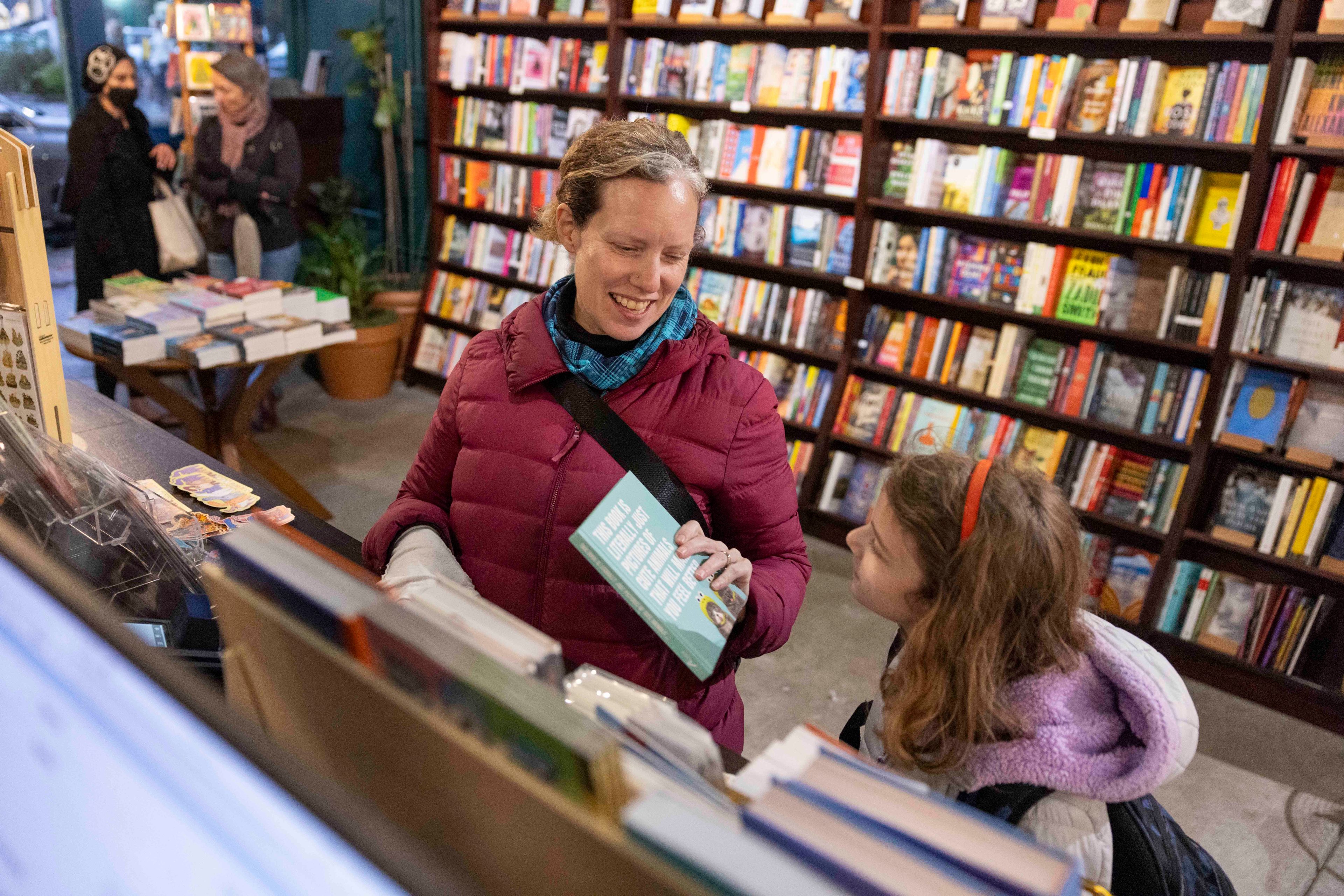 A woman in a red coat holds a book, smiling at a girl beside her. They're in a bookstore with shelves full of colorful books. Two women are in the background.