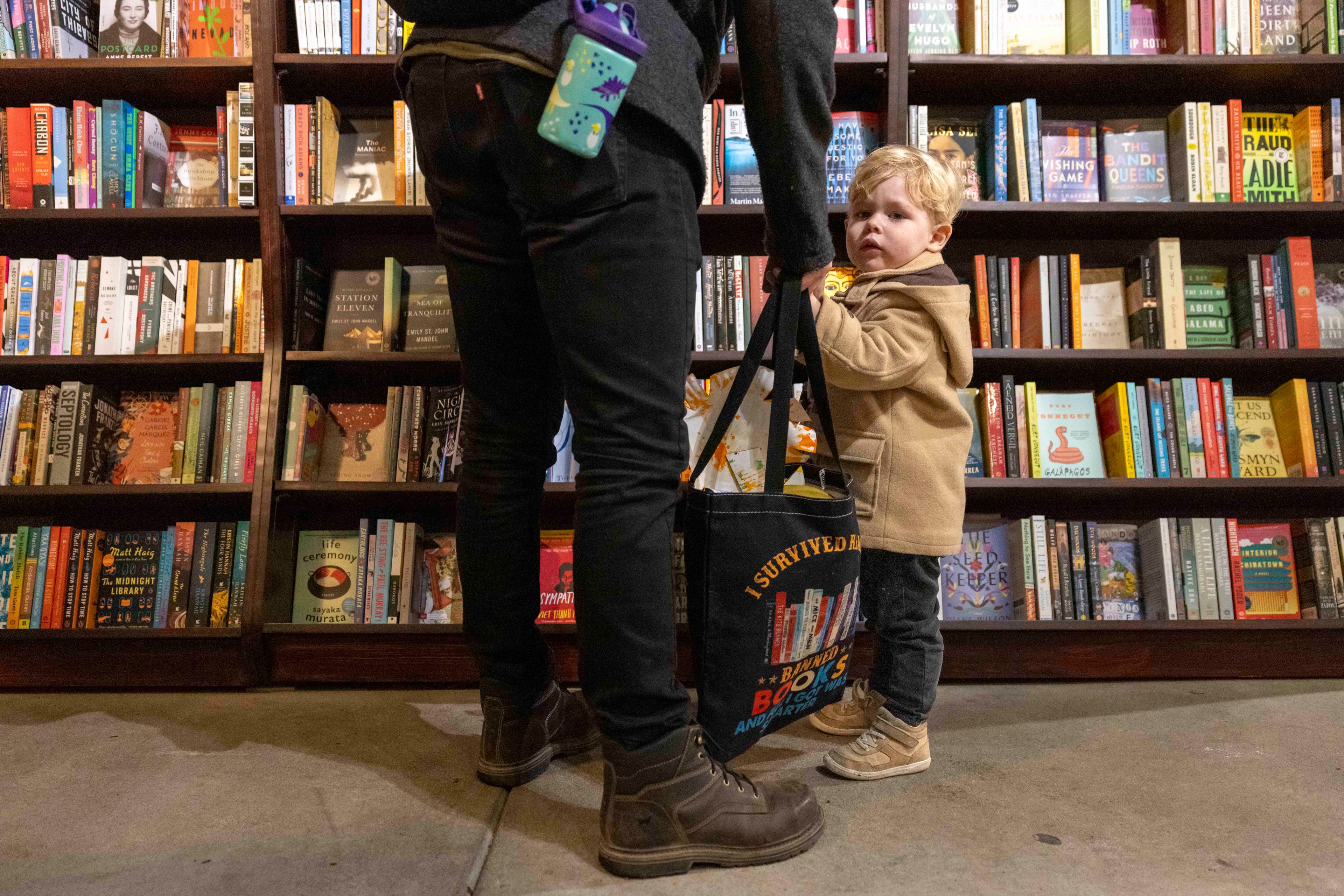 A child in a tan coat stands next to an adult holding a shopping bag in a bookstore. The shelves are full of various colorful books. The child looks curious.