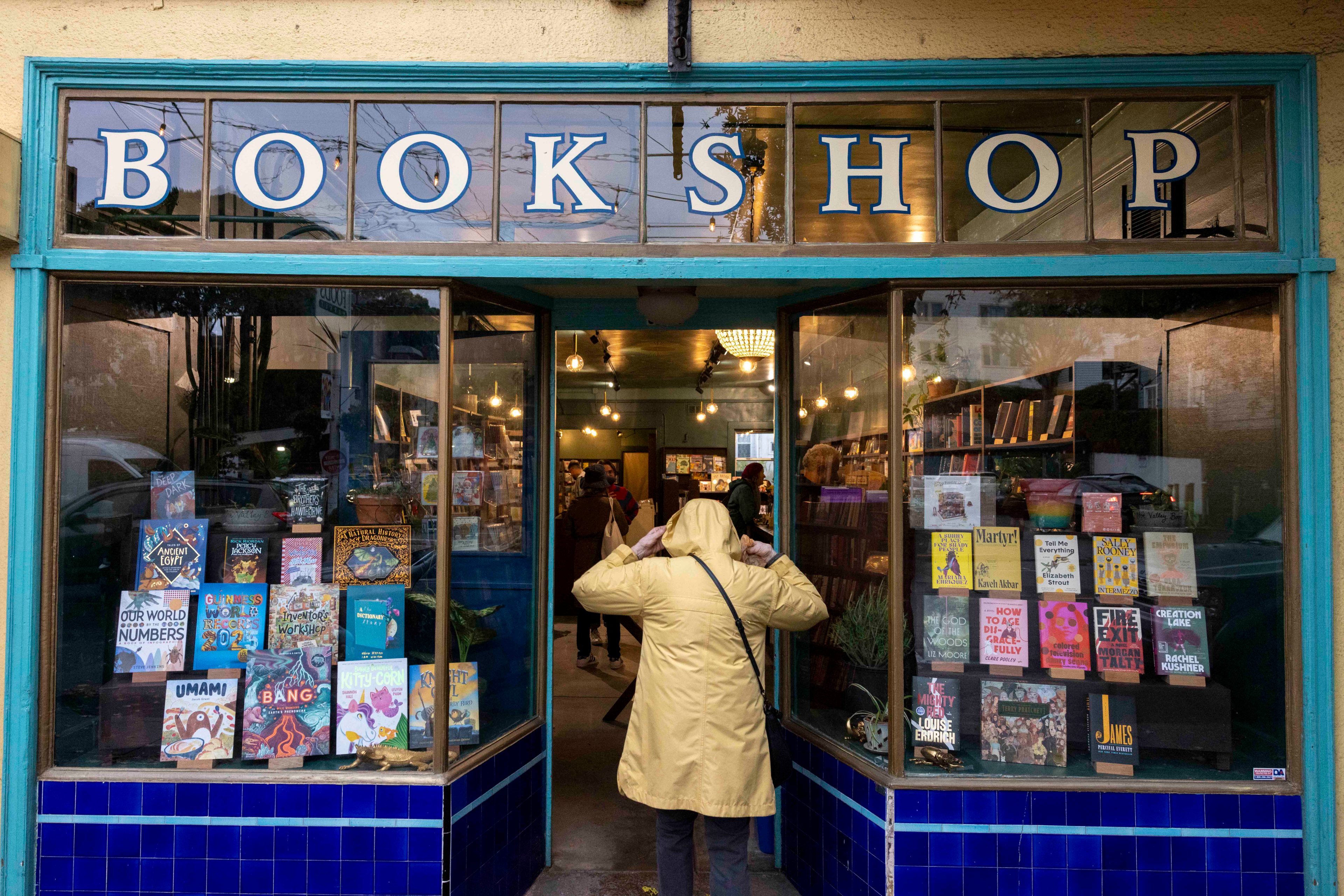 A person in a yellow coat enters a cozy bookshop with a blue exterior. The window displays various books, and warm lights illuminate the inviting interior.