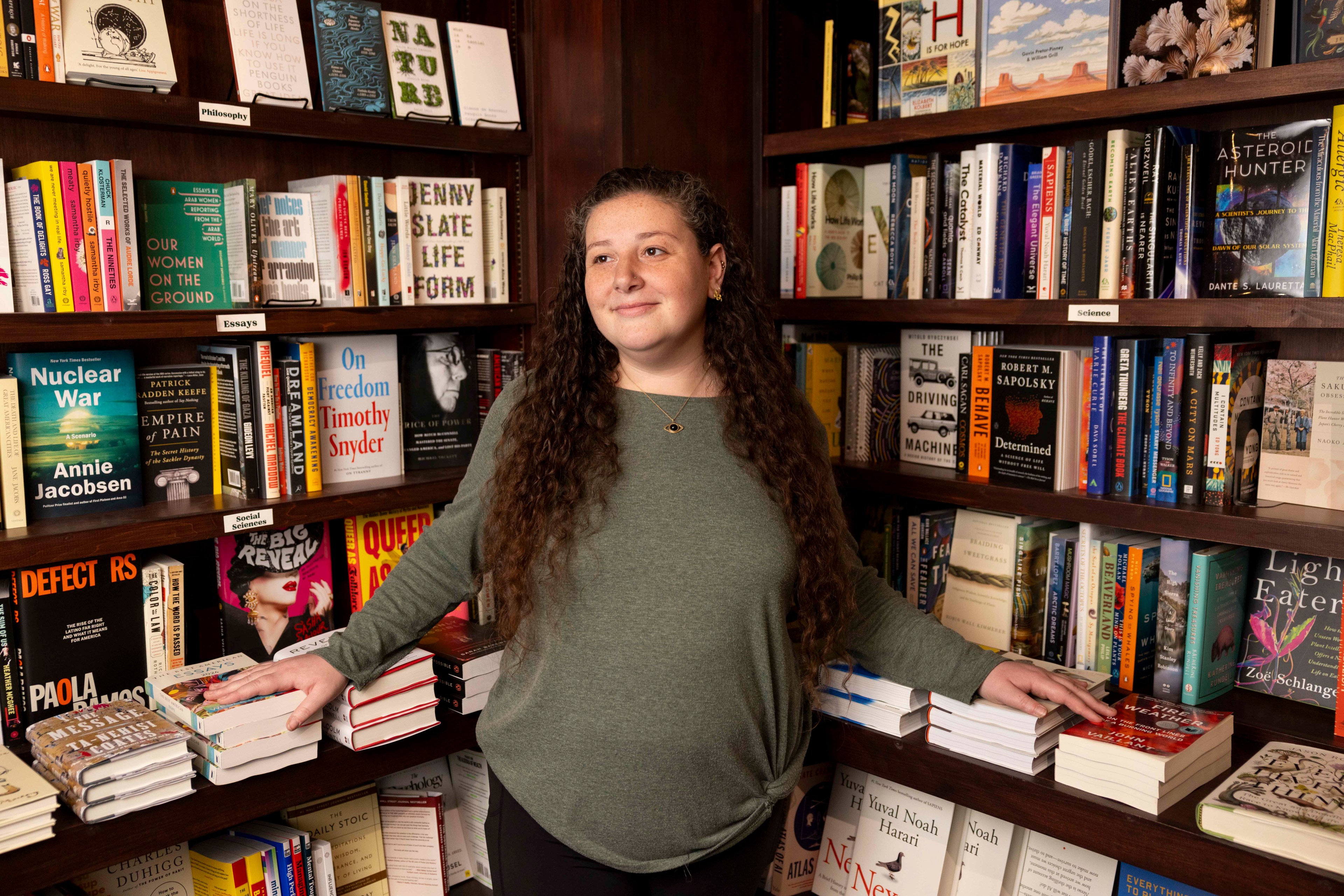 A woman with long curly hair stands in a cozy bookstore, surrounded by shelves filled with colorful books. She rests her hands on two stacks of books.