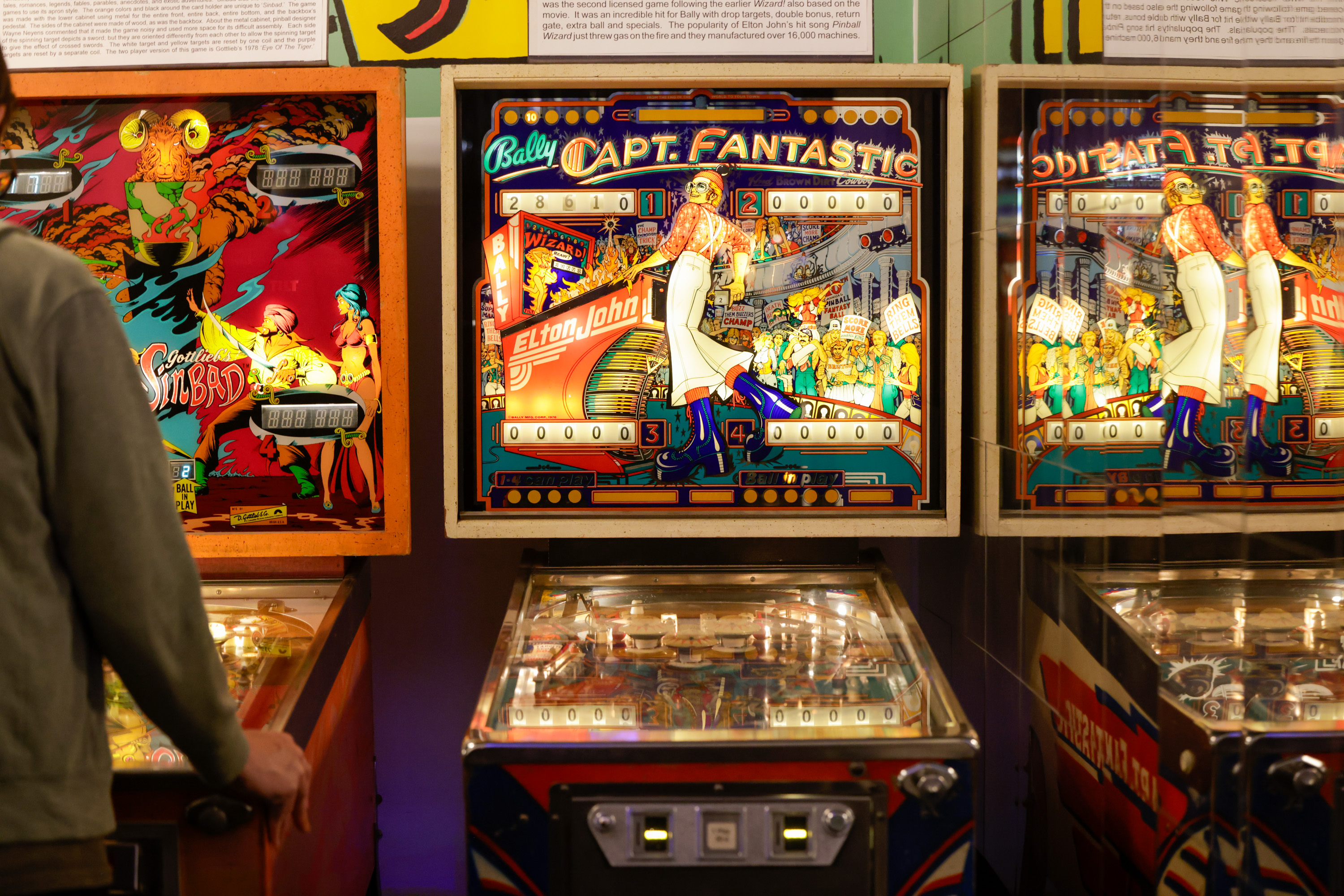 The image shows a person standing in front of two vintage pinball machines. One machine features a theme with &quot;Capt. Fantastic&quot; and the other depicts &quot;Sinbad.&quot;