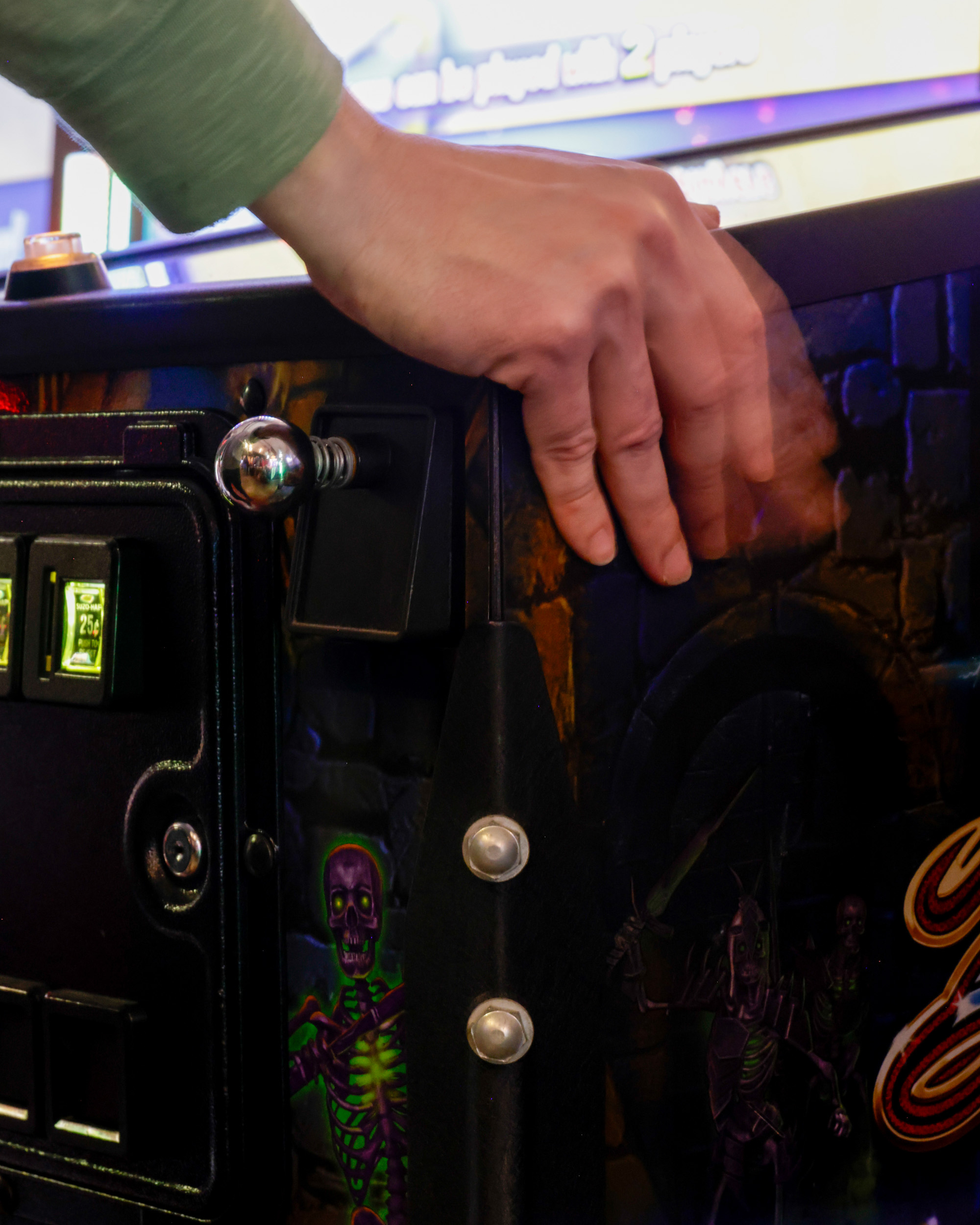 A hand pulls the lever of a pinball machine with a spooky, skeleton-themed design. The machine has coin slots labeled &quot;25¢&quot; and vibrant, colorful artwork.