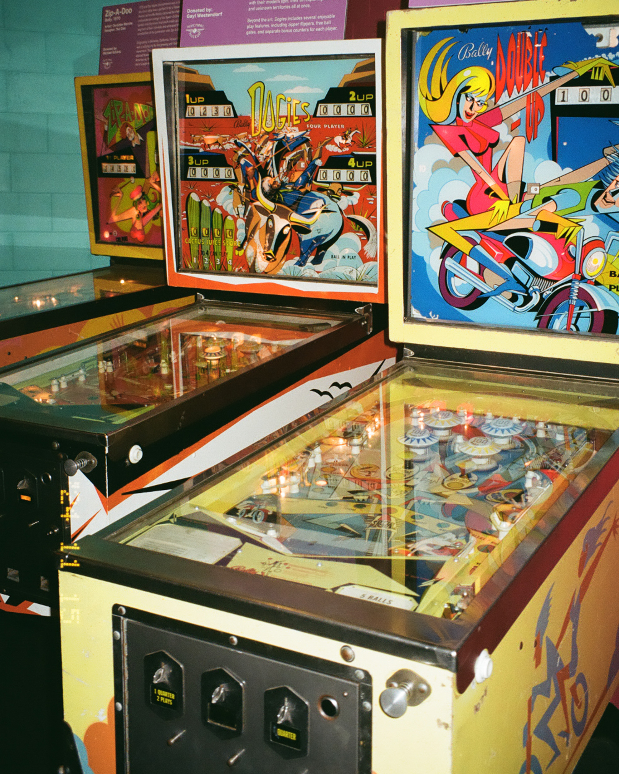 The image shows vintage pinball machines with colorful artwork, featuring cowboys and a woman on a motorbike. The machines have visible score displays.