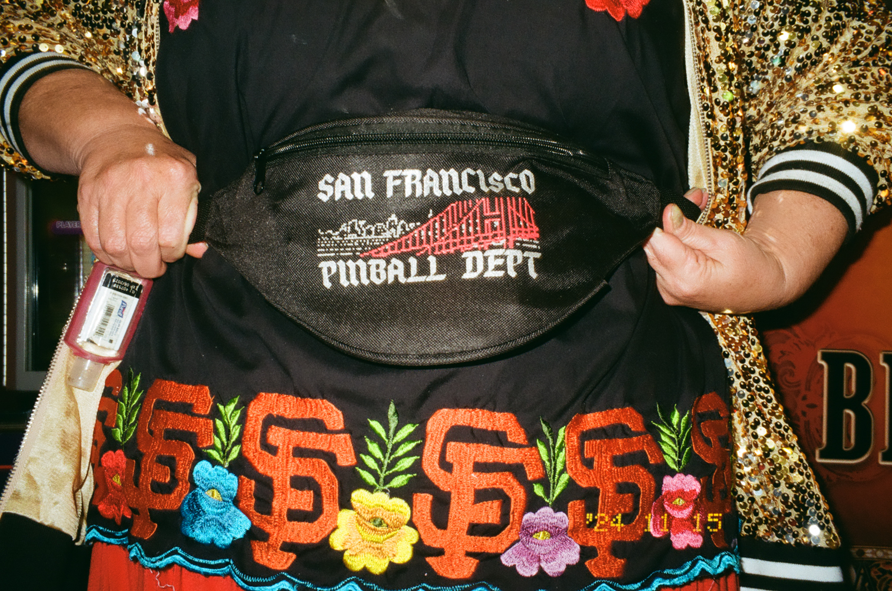 A person wears a black waist bag with &quot;San Francisco Pinball Dept&quot; text, over a jacket with gold sequins and a shirt featuring floral and SF logos.