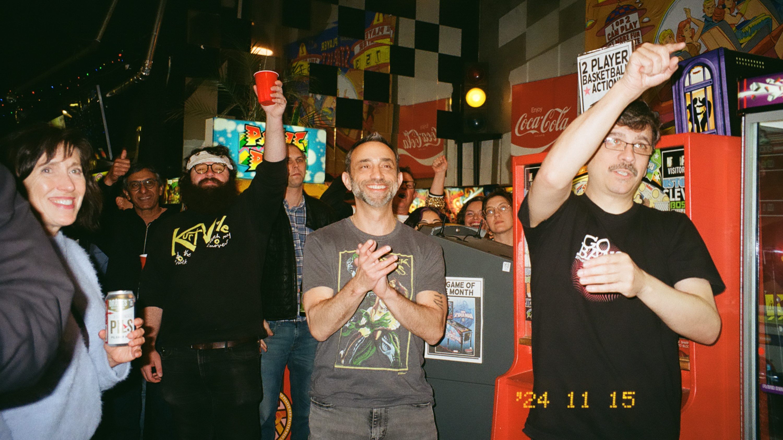 A group of people are joyfully gathered in an arcade setting, holding drinks and raising their hands in celebration. The room is colorful with arcade machines.