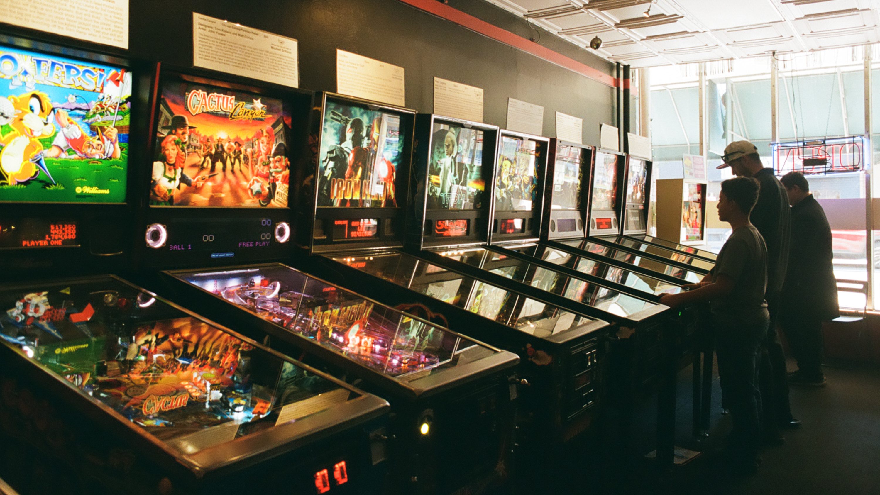 Several people play on a row of colorful pinball machines in a dimly lit arcade, with light streaming in through tall windows.