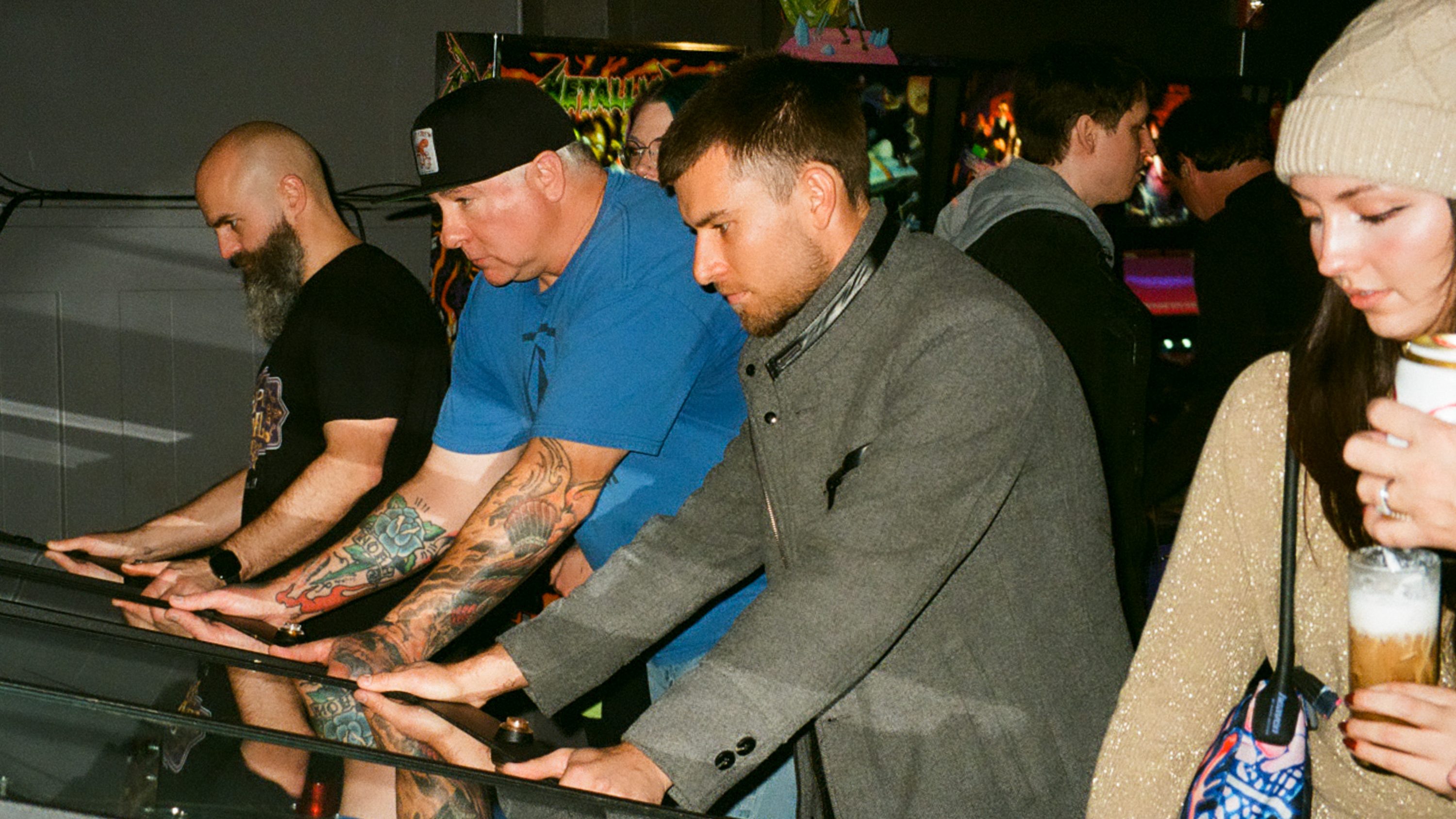 Three people are intensely playing pinball machines, while a woman nearby holds a drink, watching. The room has dim lighting, emphasizing the neon-lit machines.