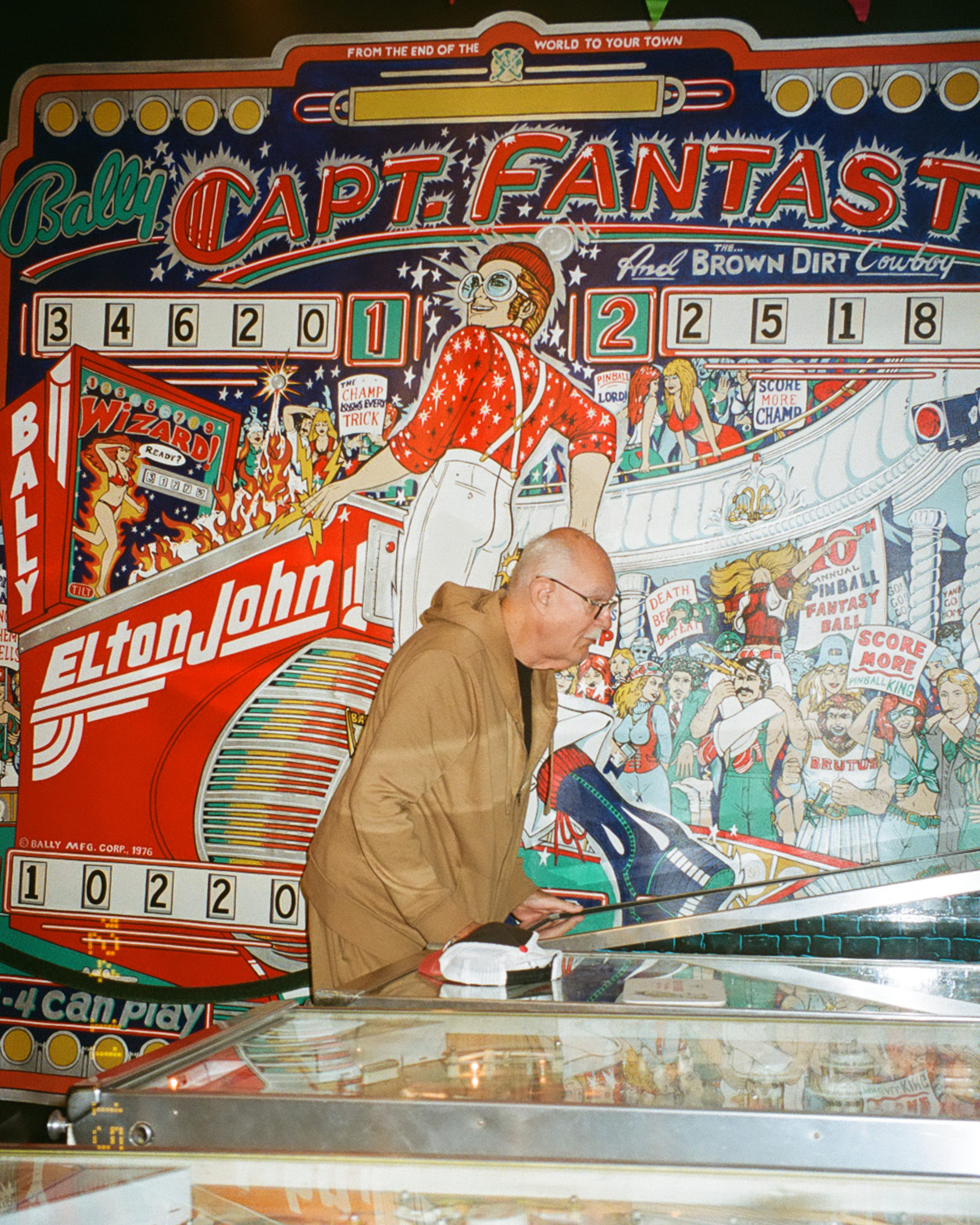 A man plays a vintage &quot;Captain Fantastic&quot; pinball machine, featuring colorful 1970s graphics with a figure resembling a rock musician and a lively crowd scene.