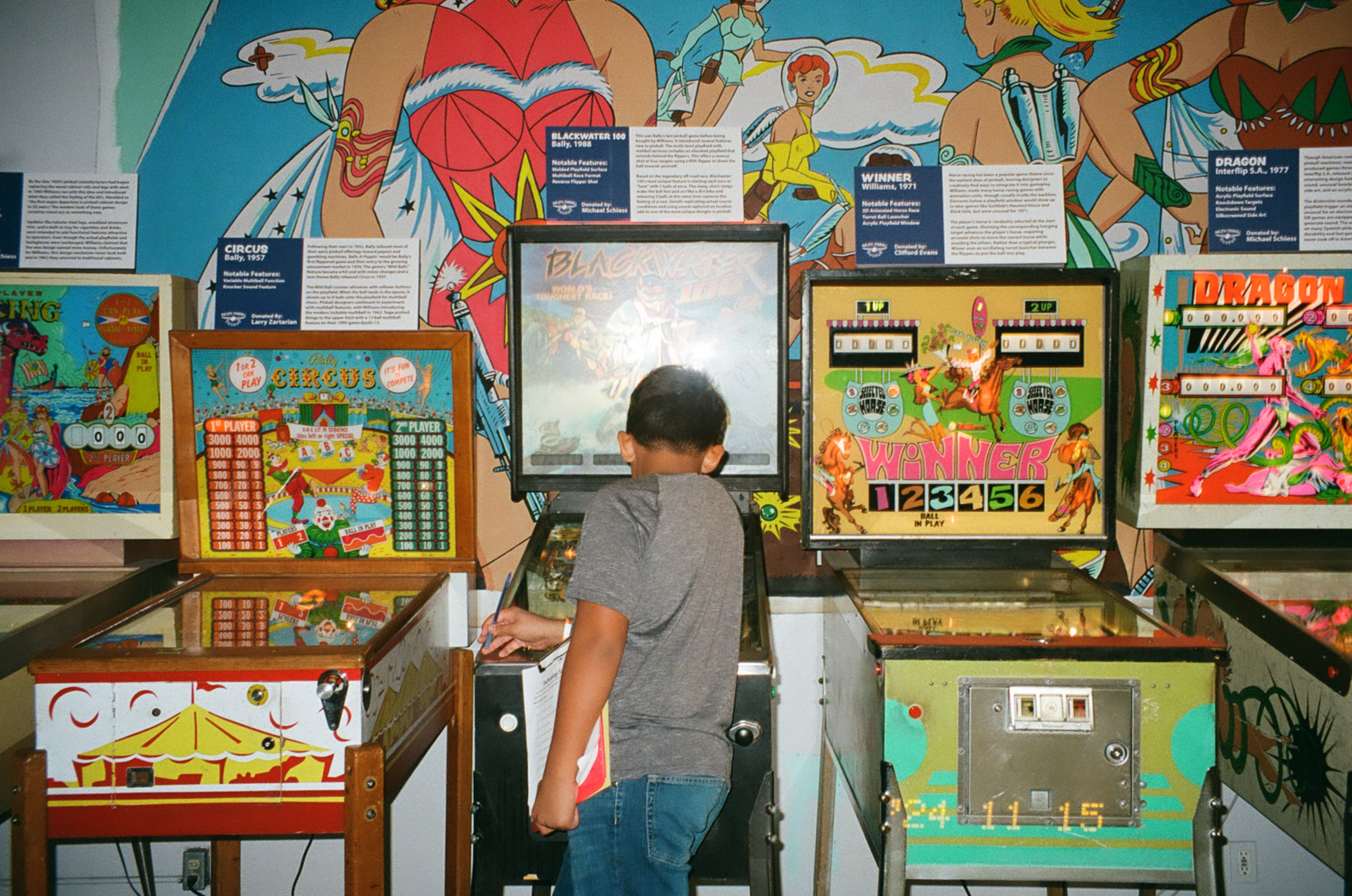 A child in a gray shirt plays vintage pinball machines, surrounded by colorful retro artwork and informational signs on a vibrant wall.